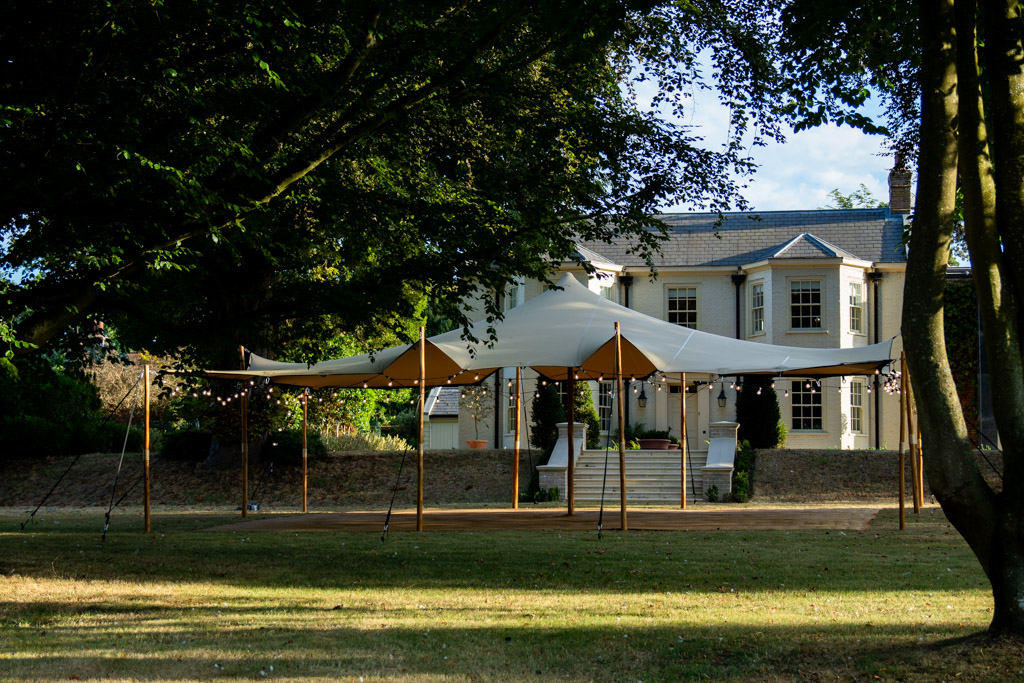 White marquee tent with string lights on a wooden lawn deck before a stately house, elegant outdoor catering setup in soft daylight.