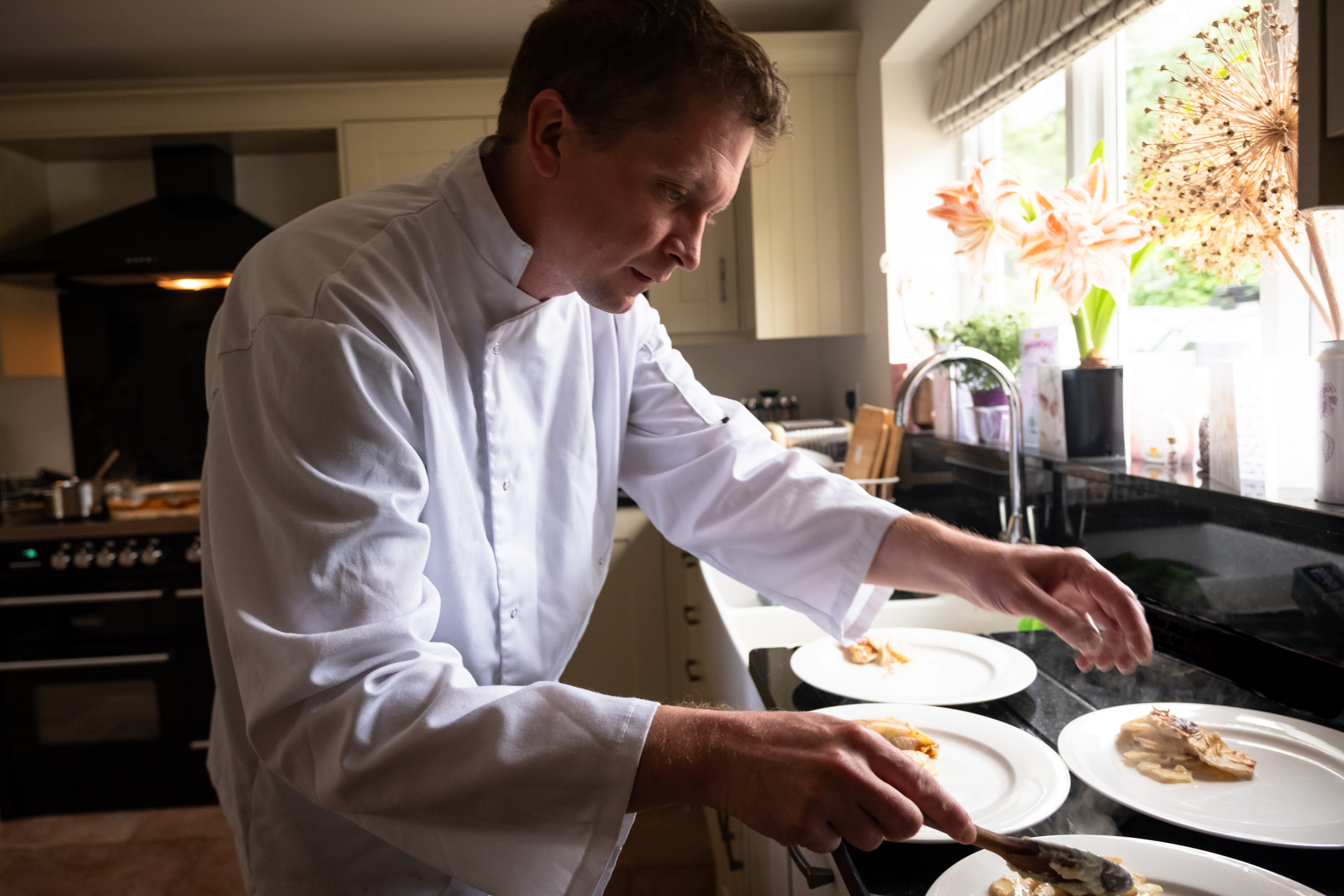 A chef in a crisp white jacket artfully plates delicate pastries on glossy white dishes amidst warm kitchen lighting and modern decor.