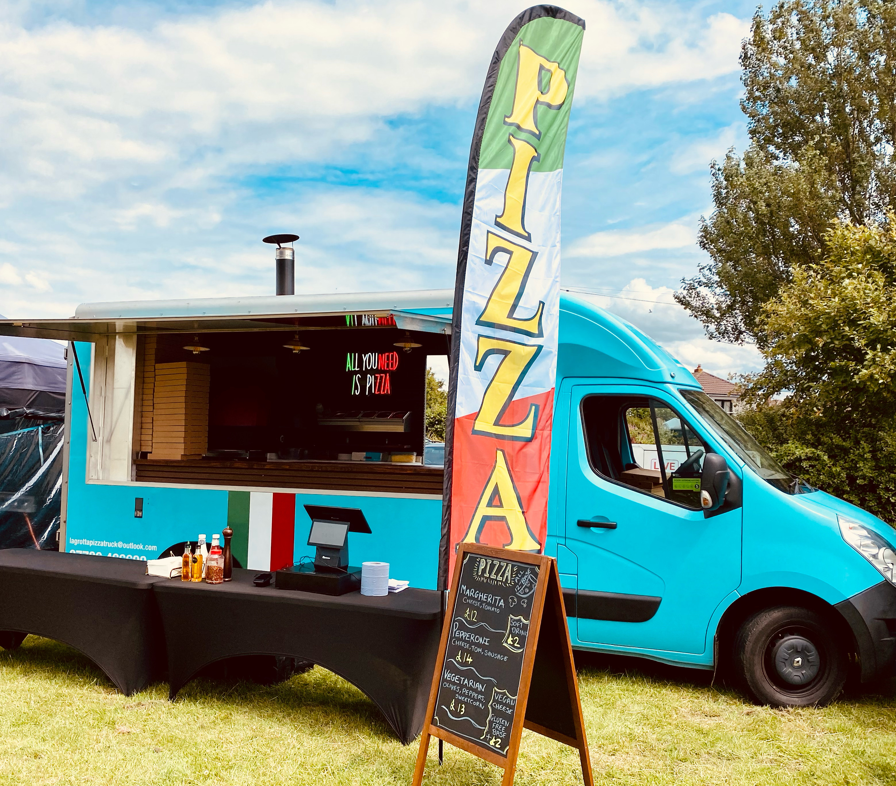 A vibrant blue pizza truck features a colorful "PIZZA" banner, offering fresh slices under warm daylight, enhancing the inviting outdoor atmosphere.