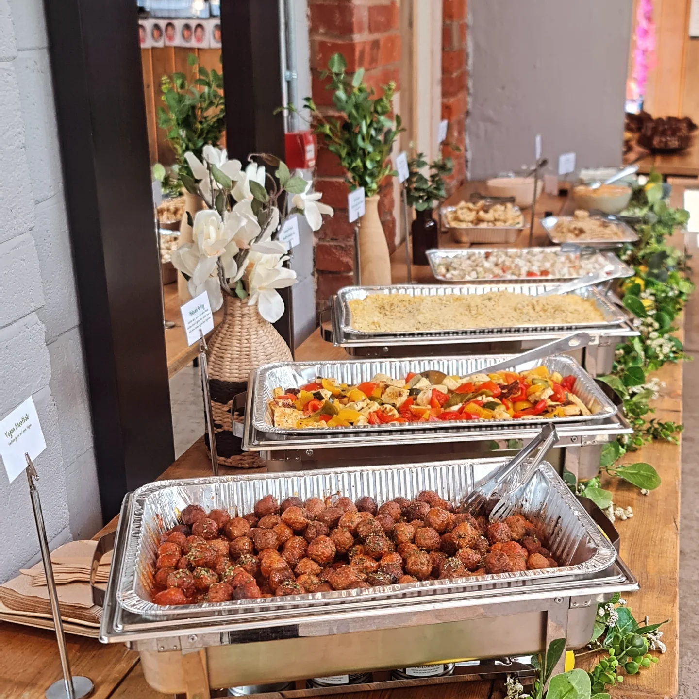 A vibrant buffet displays assorted dishes in silver trays, featuring seasoned meatballs, roasted veggies, and floral centerpieces, all under warm lighting.