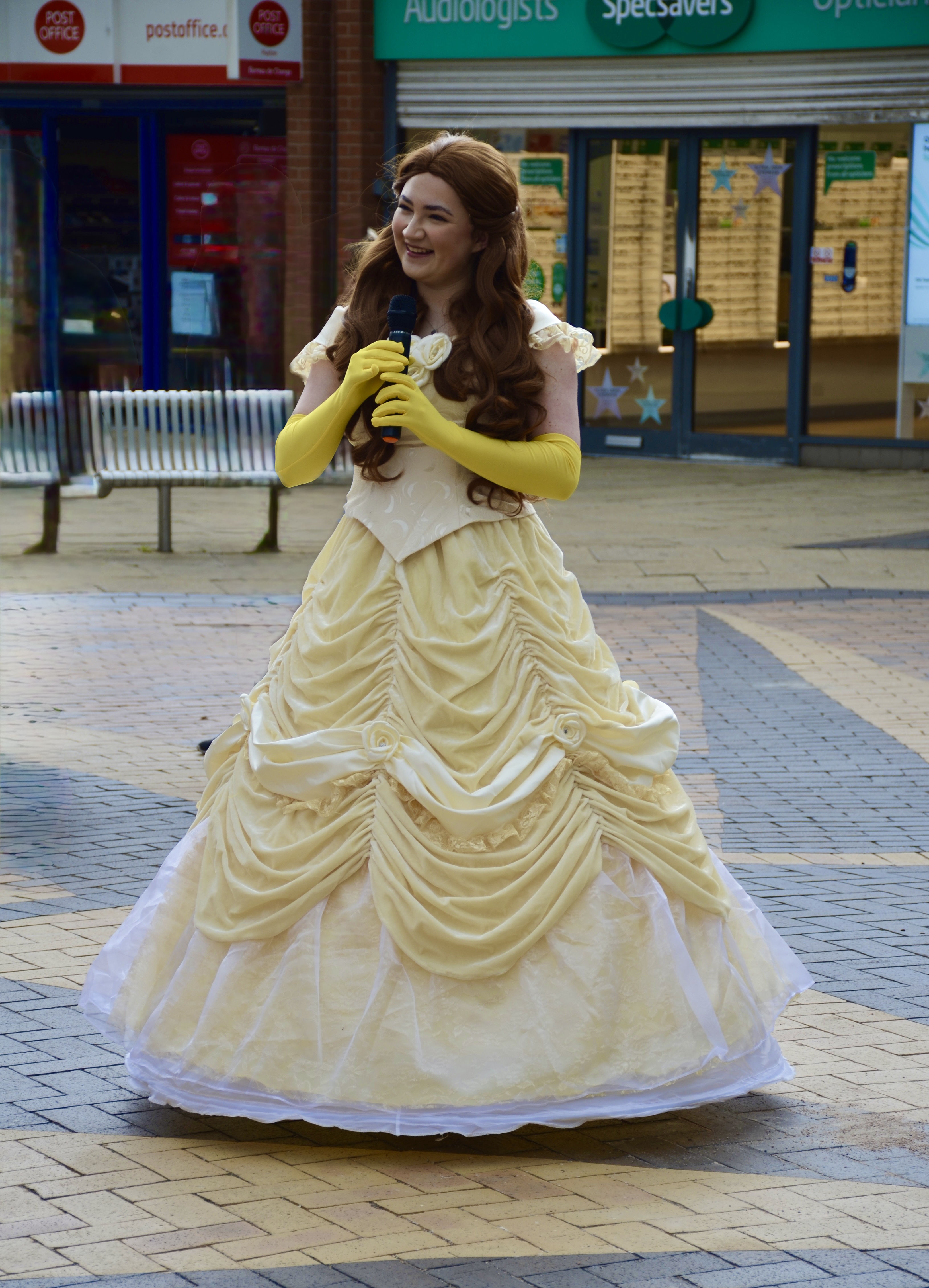 A performer in a flowing yellow gown, with intricate ruffles, stands smiling while holding a microphone in a lively outdoor shopping area.