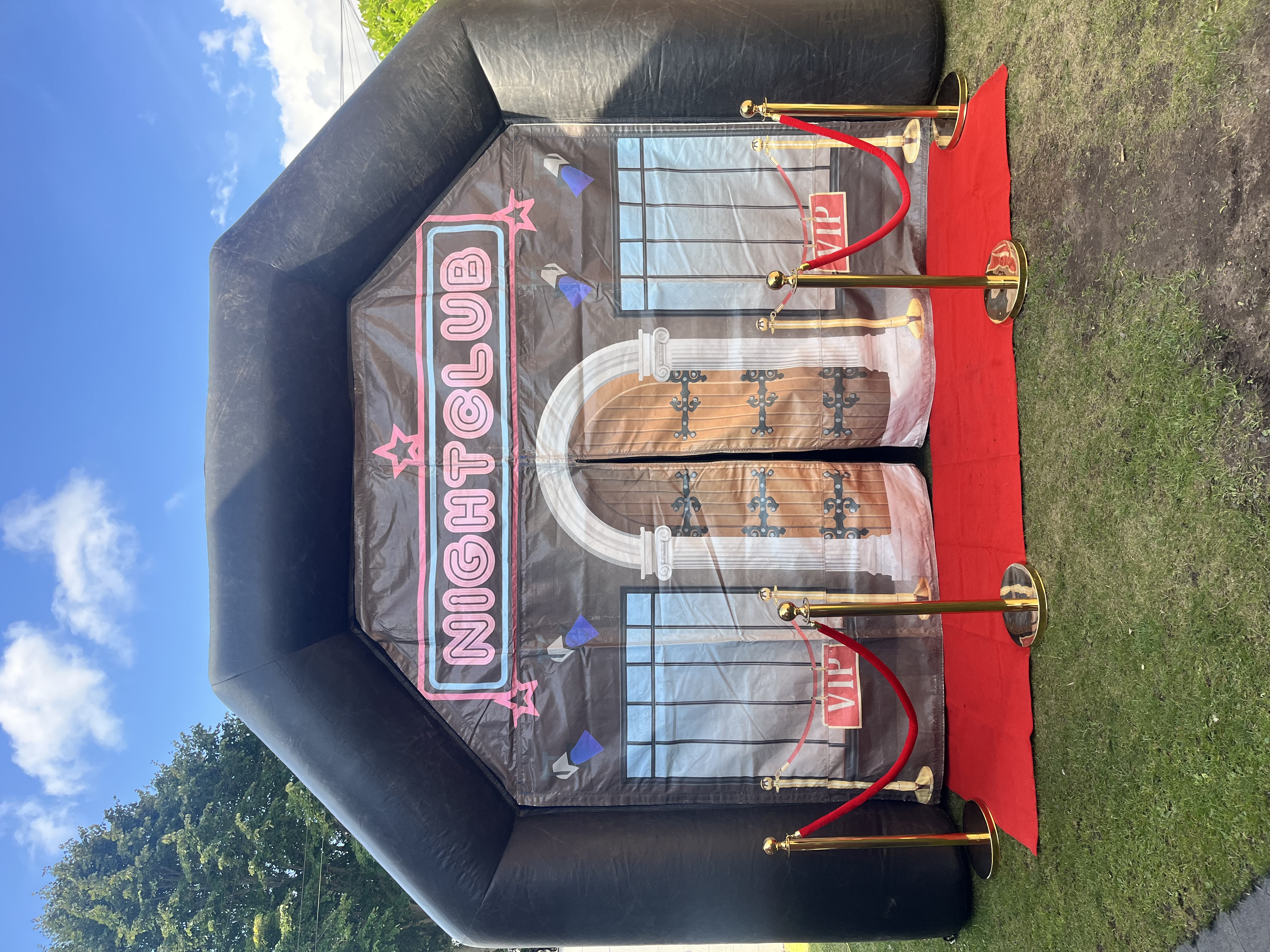 Black inflatable nightclub entrance with pink neon-style banner, red carpet, gold stanchions, and bright daylight event branding.
