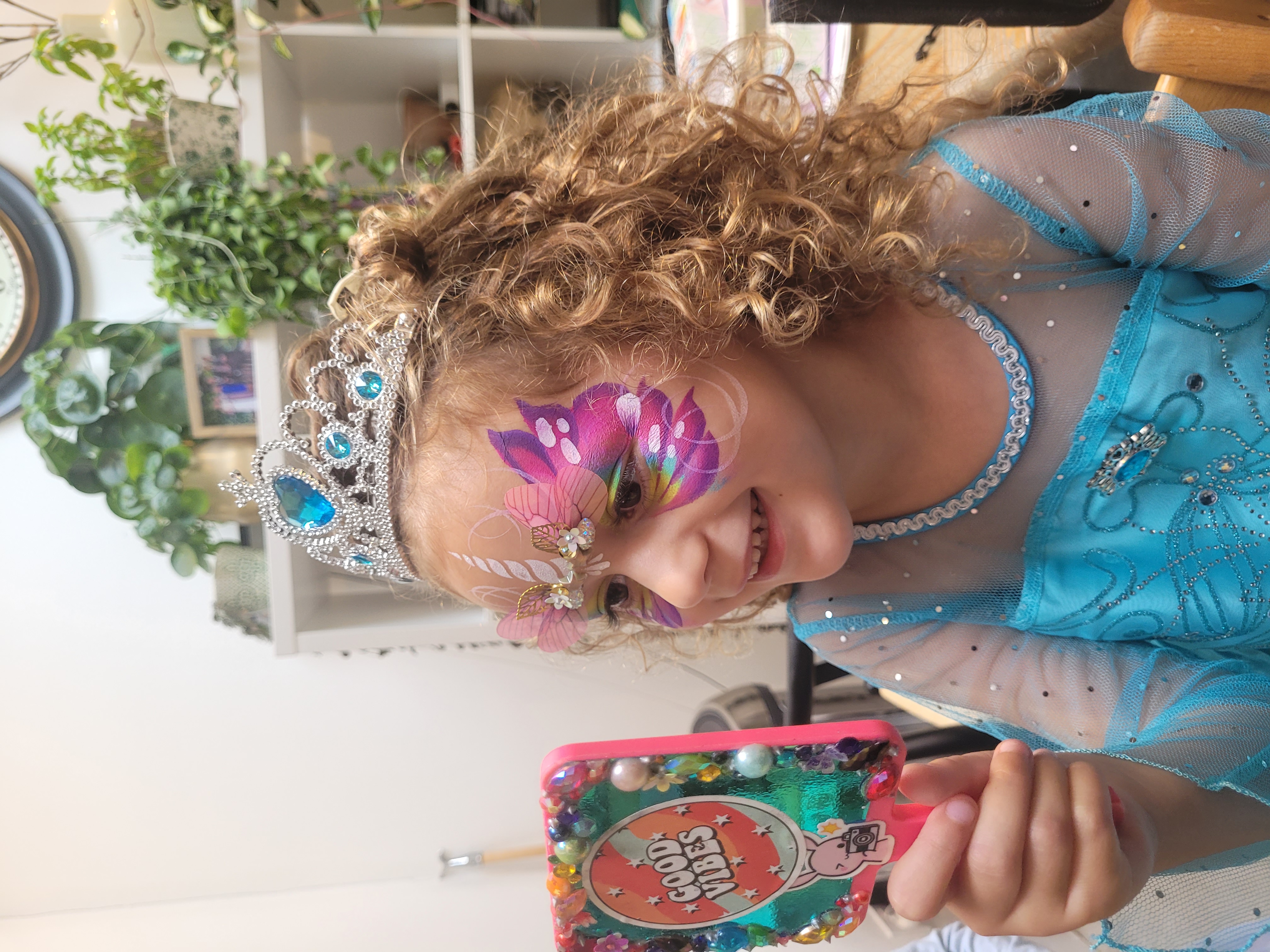 A young girl in a sparkling blue dress and tiara admires her colorful butterfly face paint in a bright, cheerful space filled with plants.