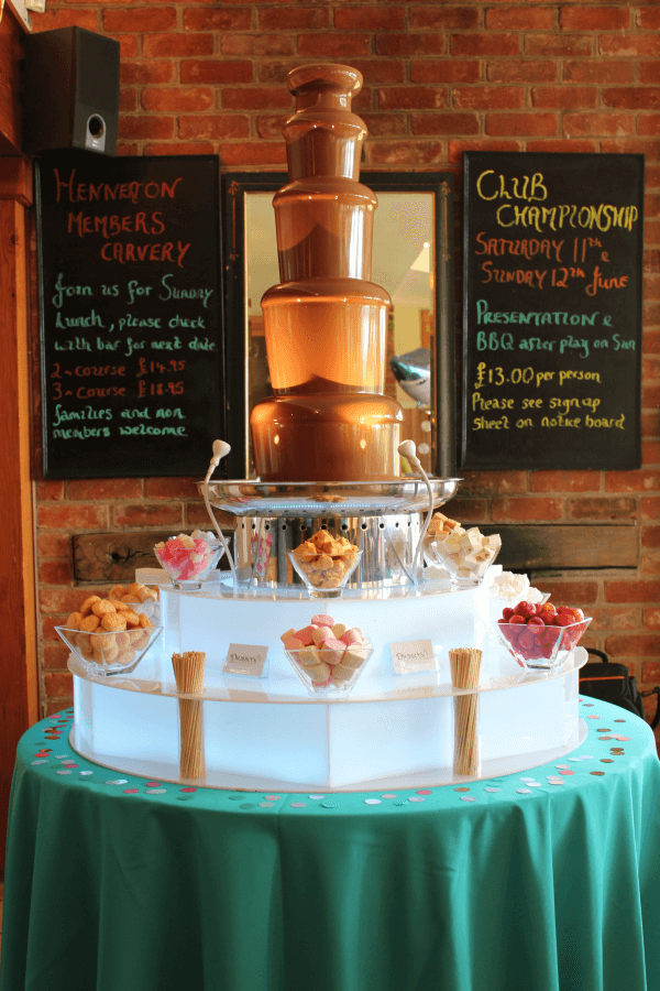 A gleaming chocolate fountain stands prominently, surrounded by plates of popcorn and grapes, with guests enjoying a festive, cheerful atmosphere.