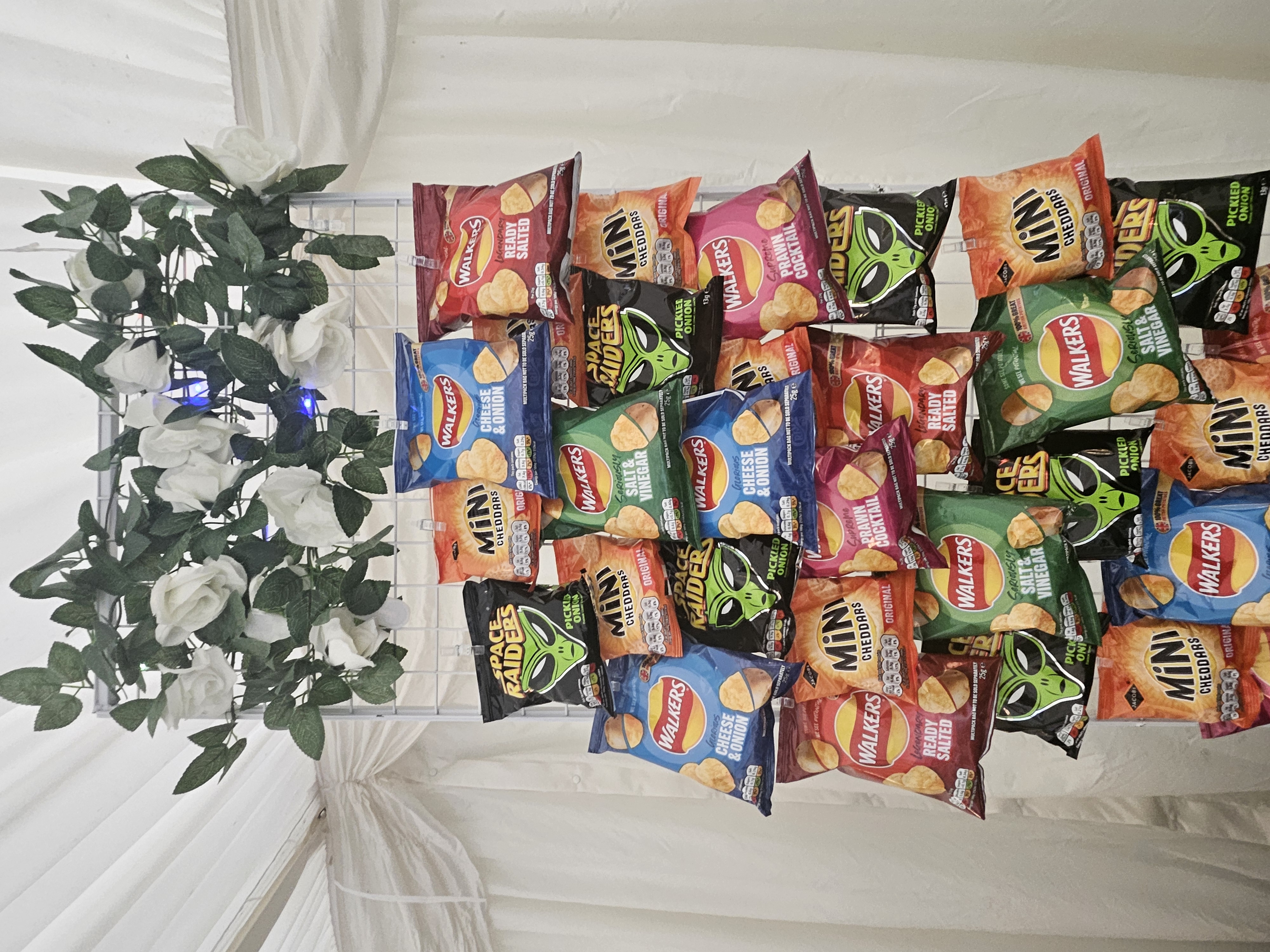Crisp catering display of Walkers crisps and Mini Cheddars on a white metal grid, topped with faux roses under soft tent lighting.