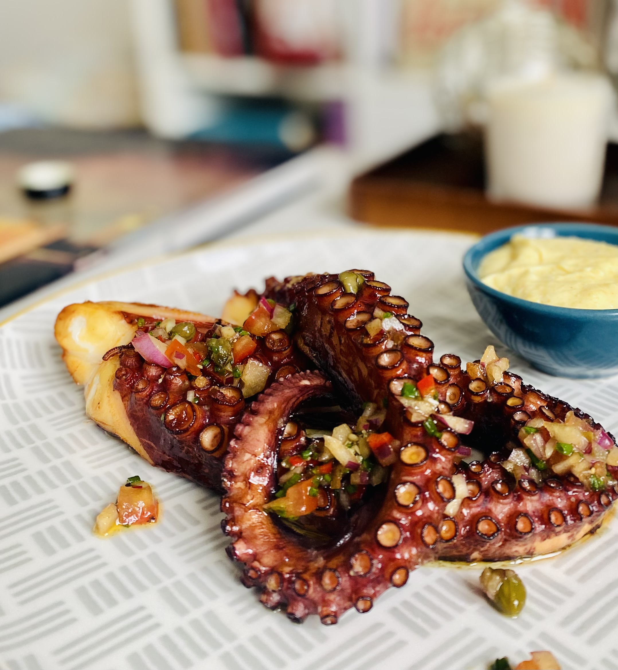 Grilled octopus, garnished with fresh salsa, rests on a patterned plate next to a blue bowl of creamy sauce, creating a vibrant, inviting display.