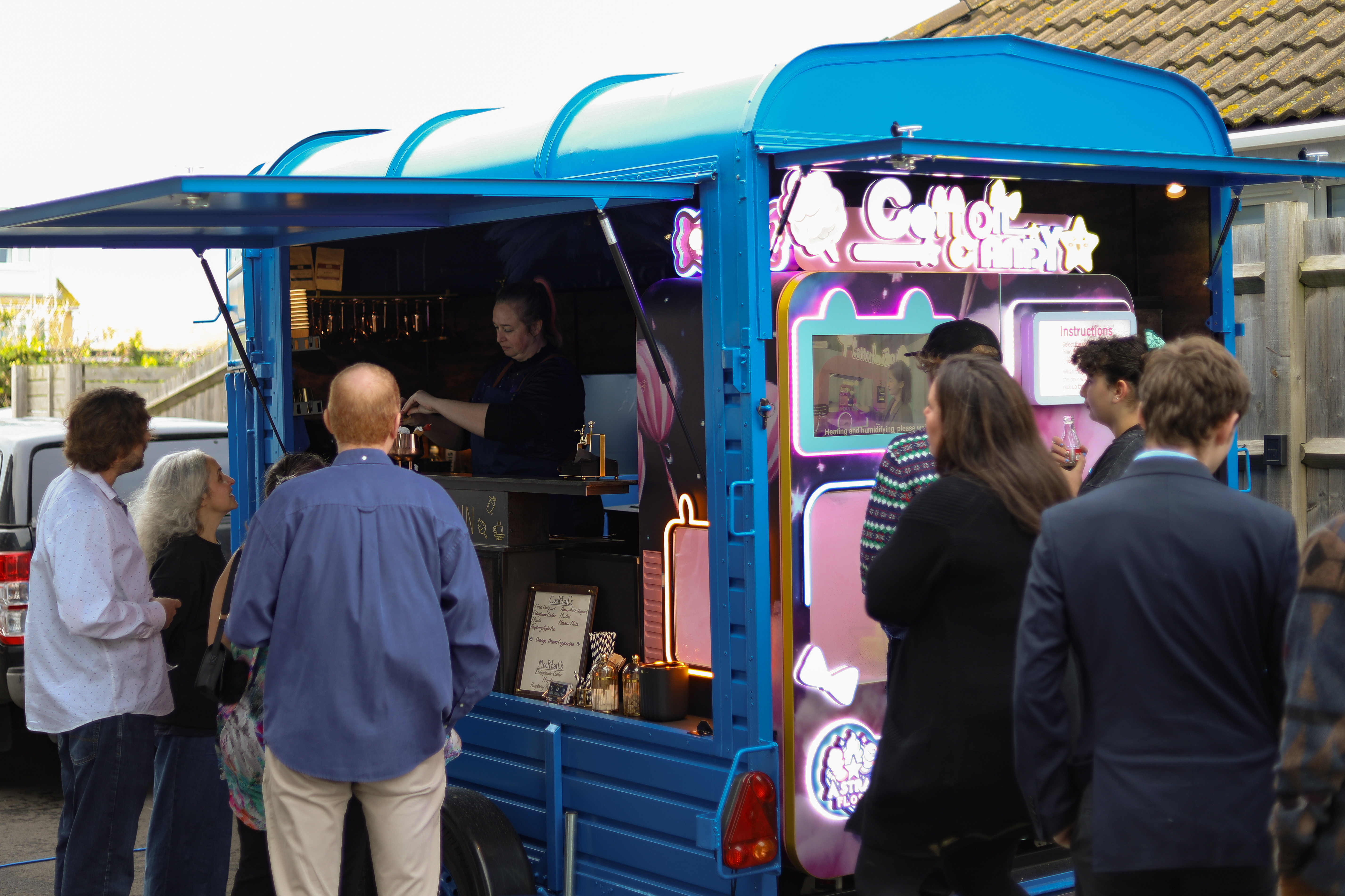 A vibrant blue vintage van serves cotton candy, adorned with neon lights. Guests gather excitedly, enjoying the festive atmosphere in soft daylight.