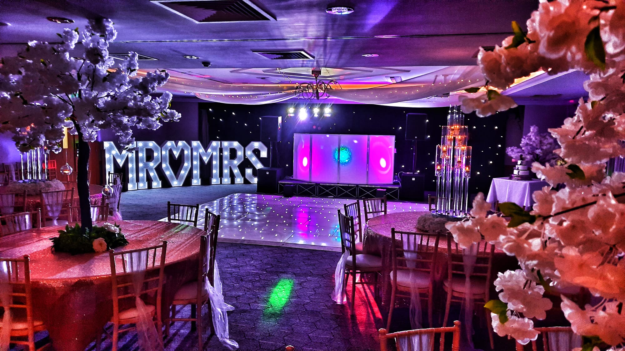 Purple-lit wedding reception with glowing “MOMRS” sign, mirrored dance floor, gold chairs, floral centerpieces, and polished buffet-style staging.