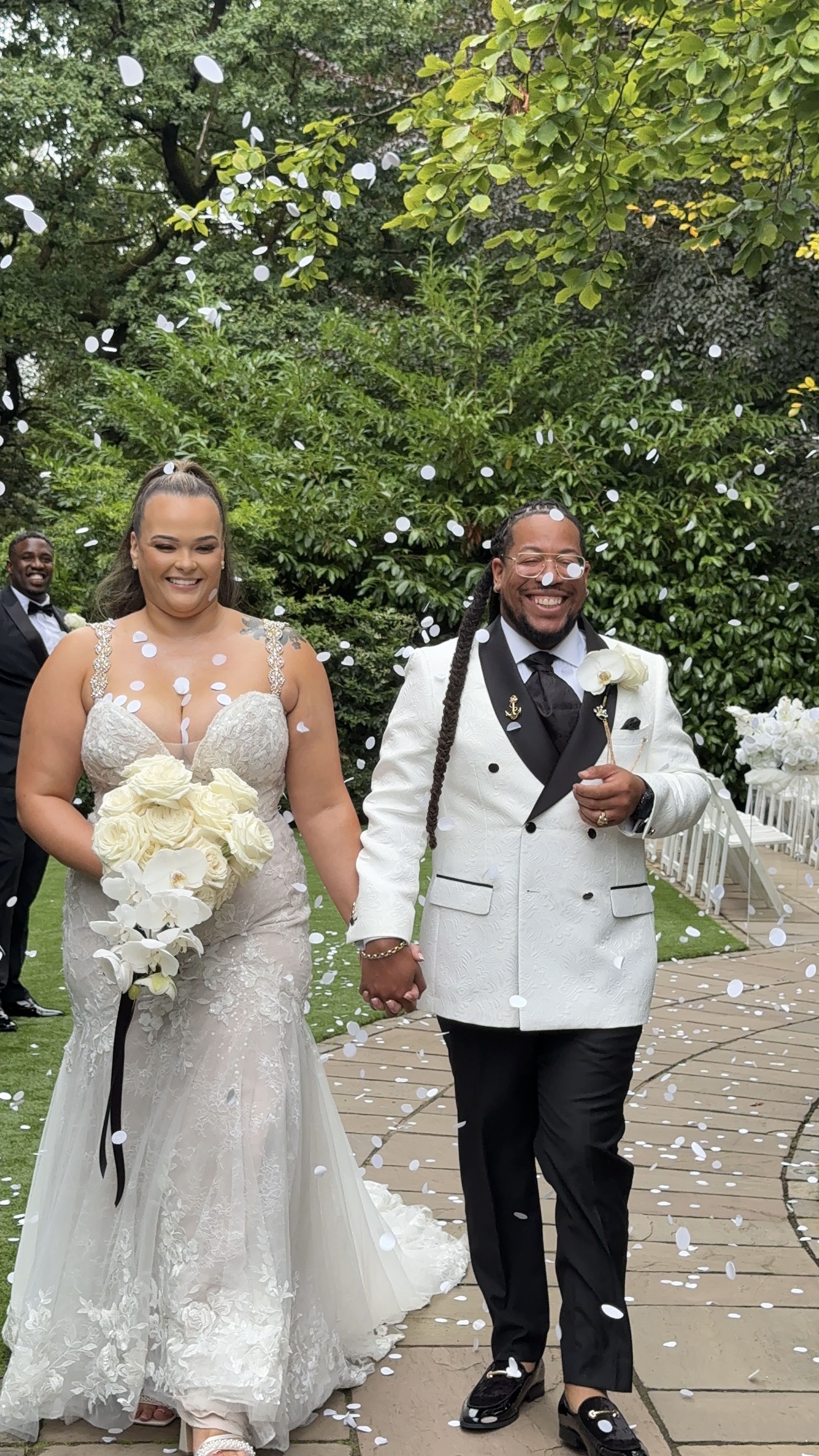 Joyful outdoor wedding ceremony with confetti, a bride in lace and groom in white tux stroll hand-in-hand on a garden path.