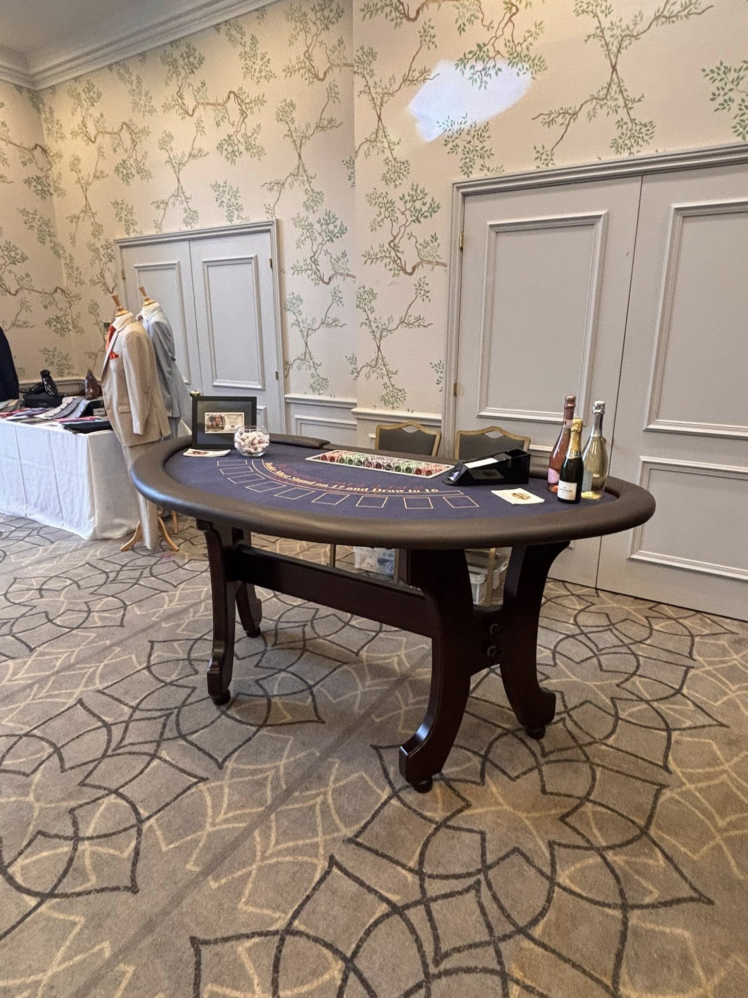 A polished casino table with a felt surface is elegantly placed against a floral wallpaper, complemented by champagne bottles and suits.