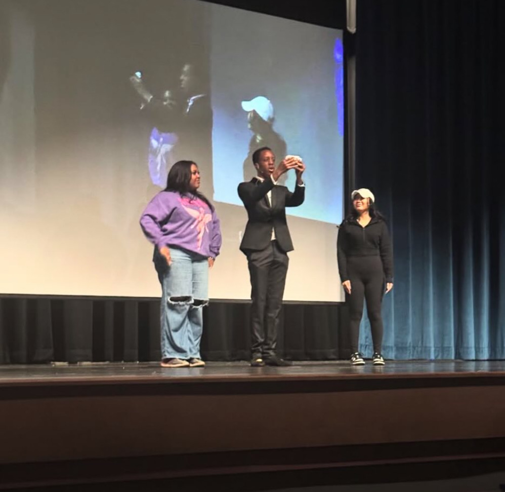 Three presenters stand on a lit stage before a projection screen, one filming a selfie, in a casual live event with theater curtains.