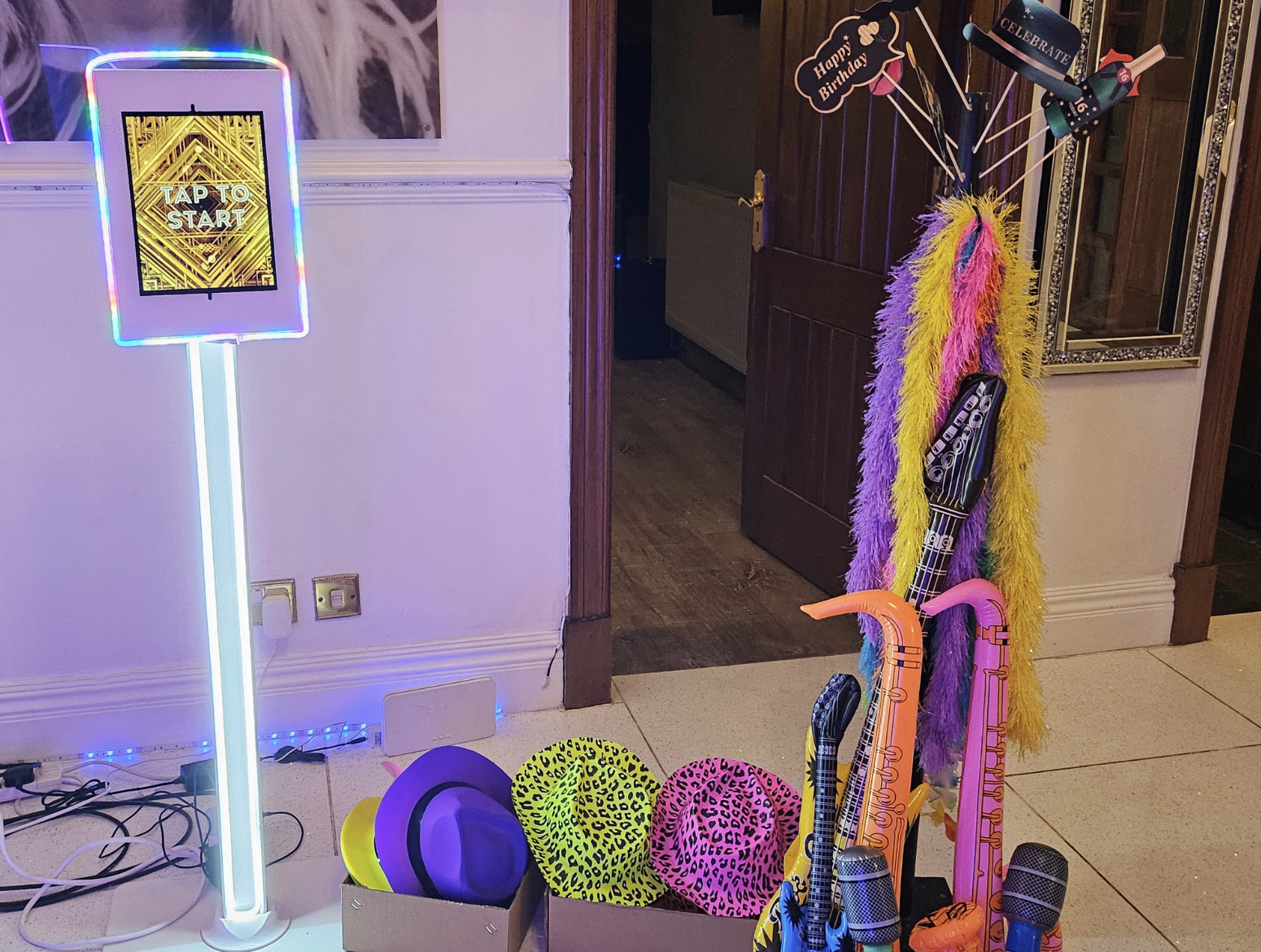 A vibrant selfie pod features a lit-up mirror and playful props, including colorful hats and feather boas, set against a cozy backdrop.
