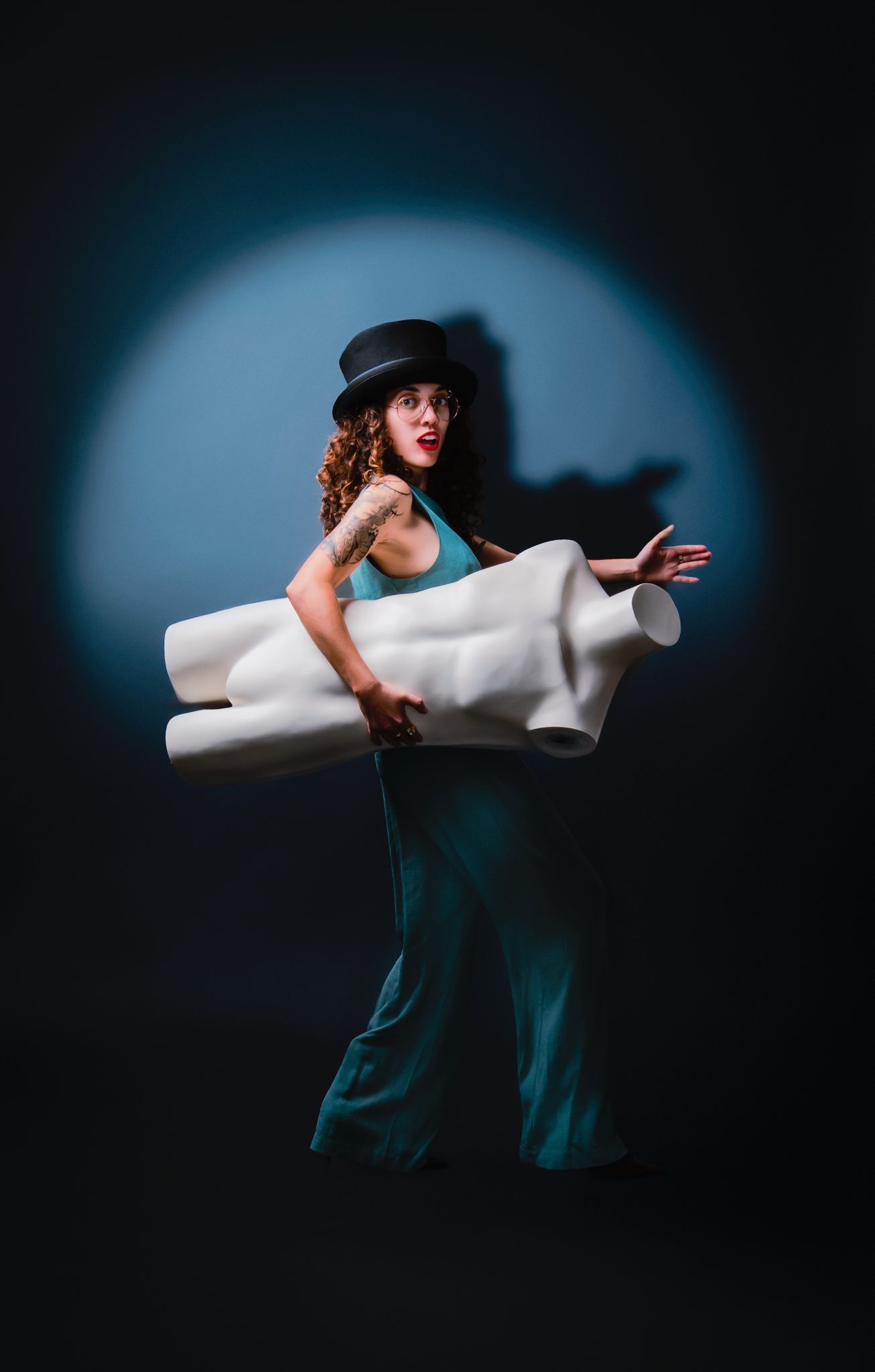Spotlit performer in teal jumpsuit and top hat carries a white sculptural prop, with dramatic studio lighting and sleek, artistic vibe.