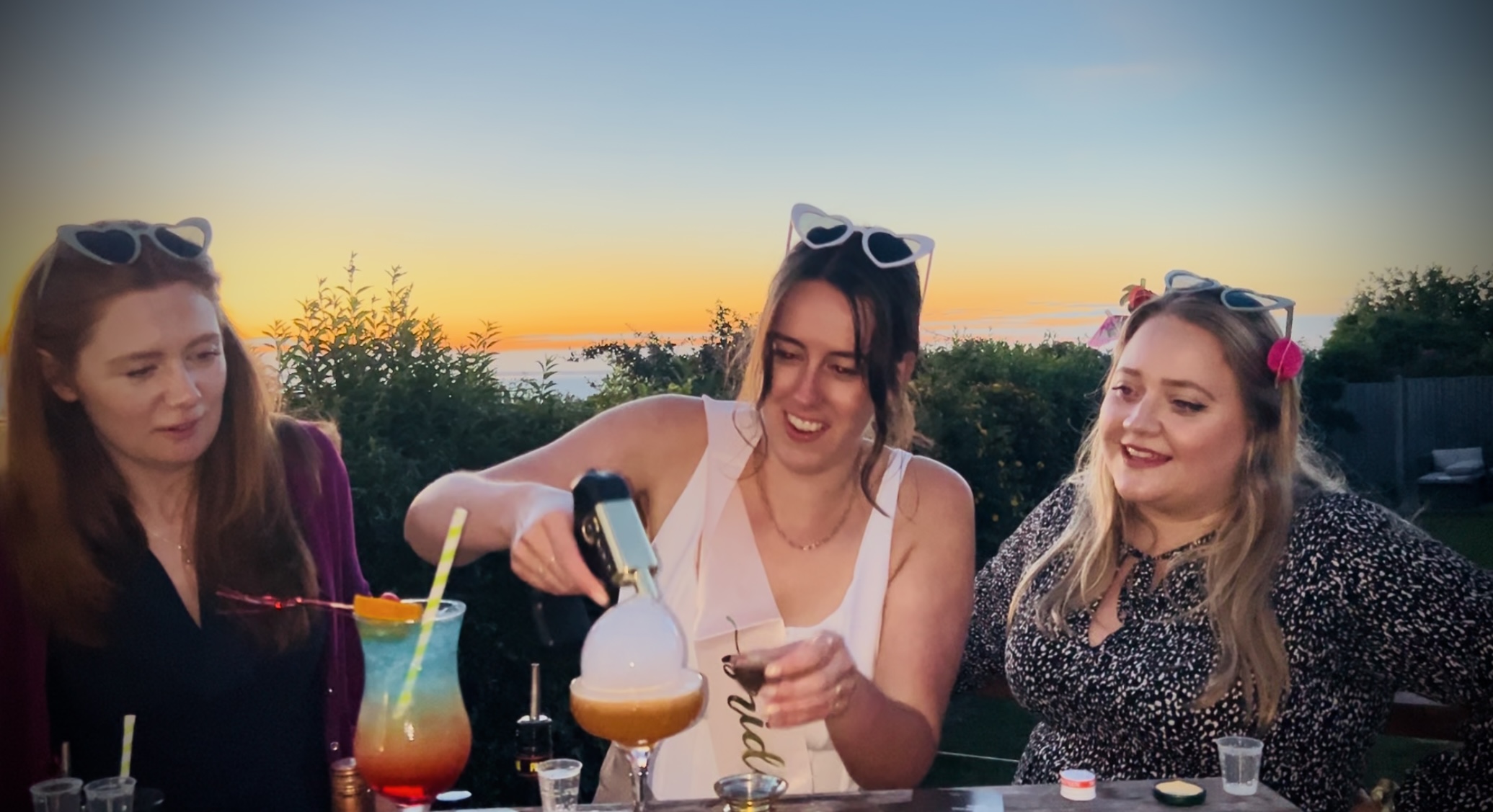 Three friends are joyfully crafting colorful cocktails at sunset, surrounded by lush greenery and a vibrant atmosphere filled with laughter.