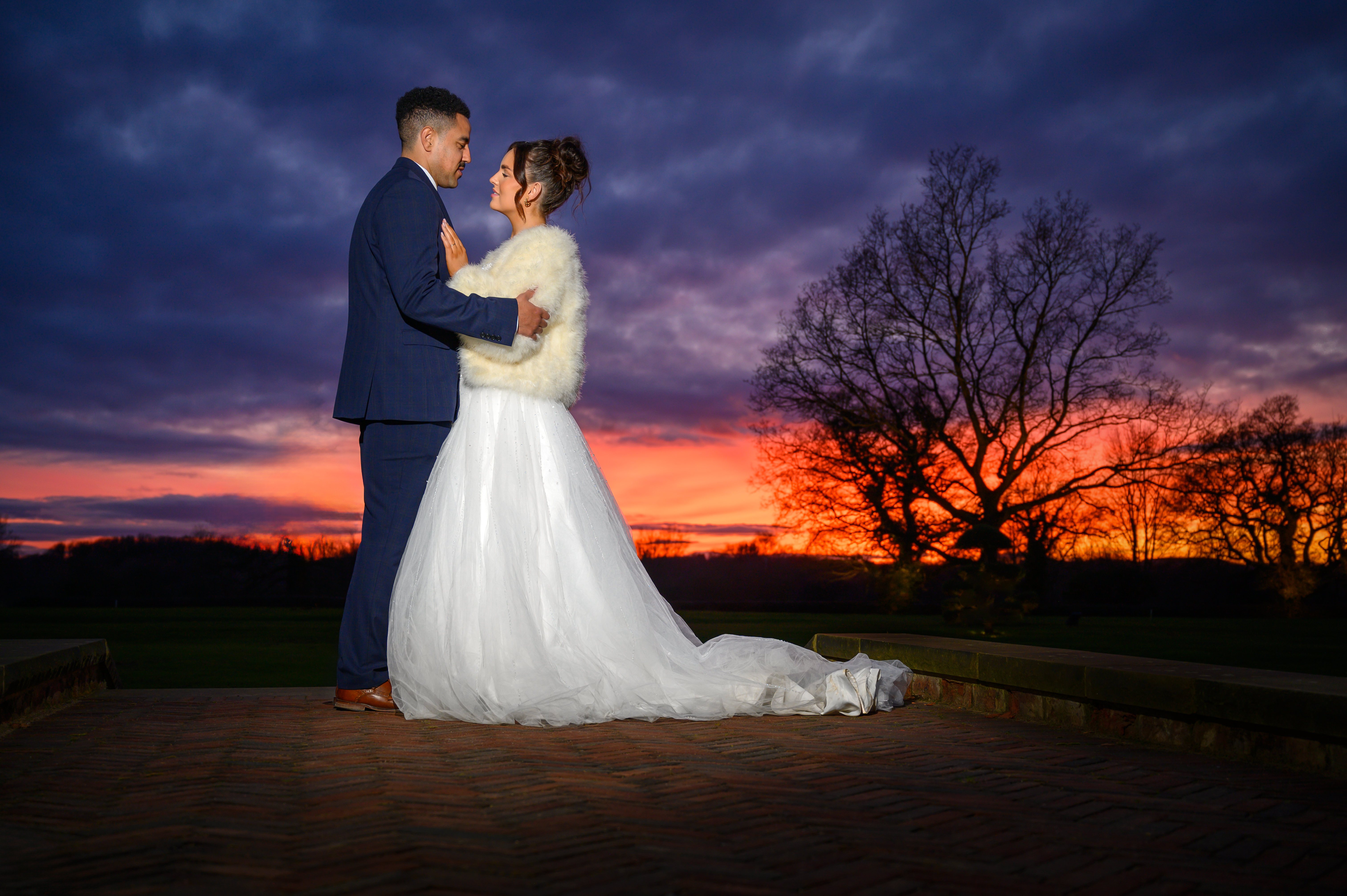 A couple in elegant attire shares a tender moment against a vibrant sunset, creating a romantic atmosphere with soft, warm hues.