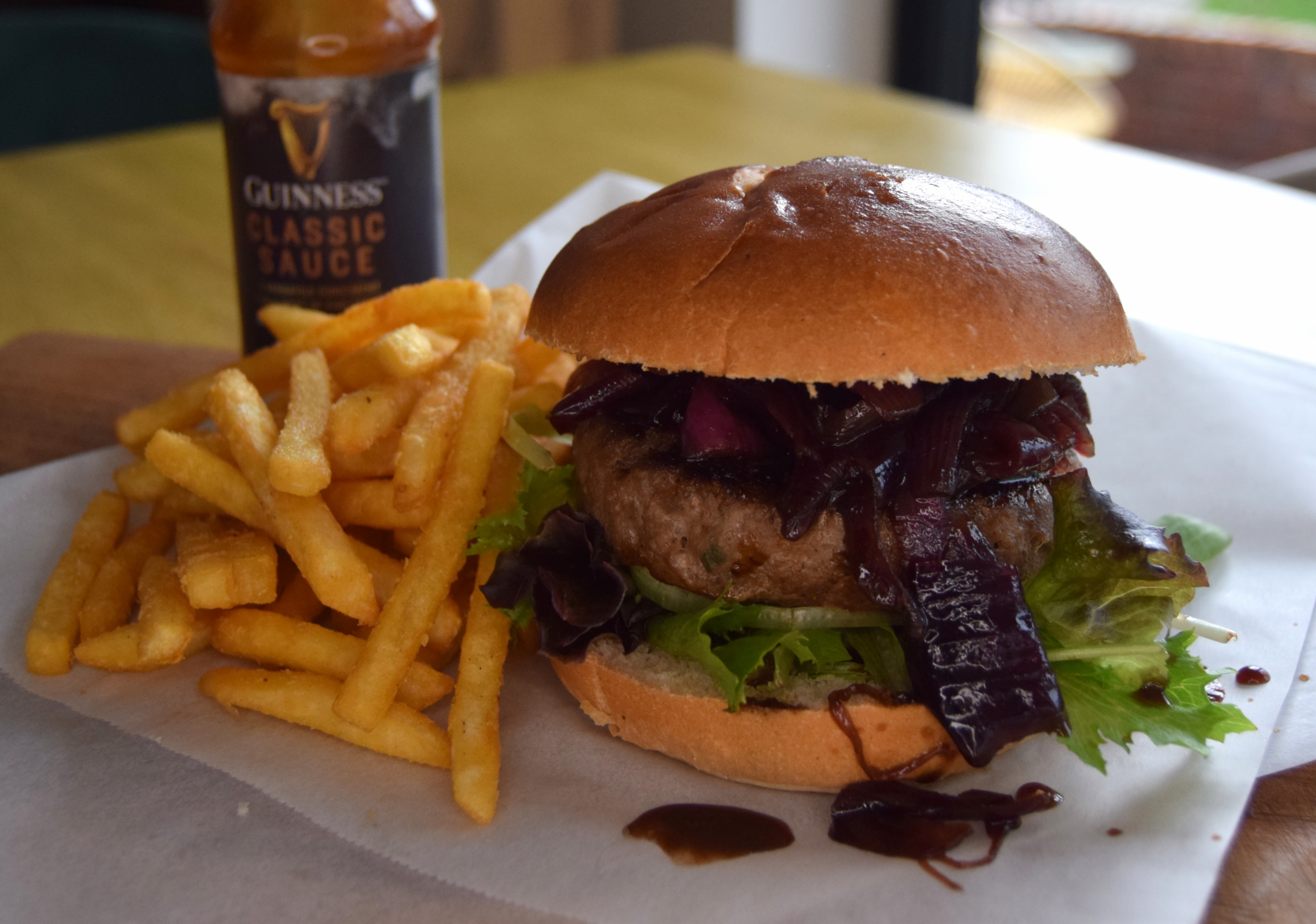 A succulent burger topped with grilled onions and fresh lettuce sits beside crispy fries, complemented by a bottle of Guinness sauce, all on a rustic wooden board.