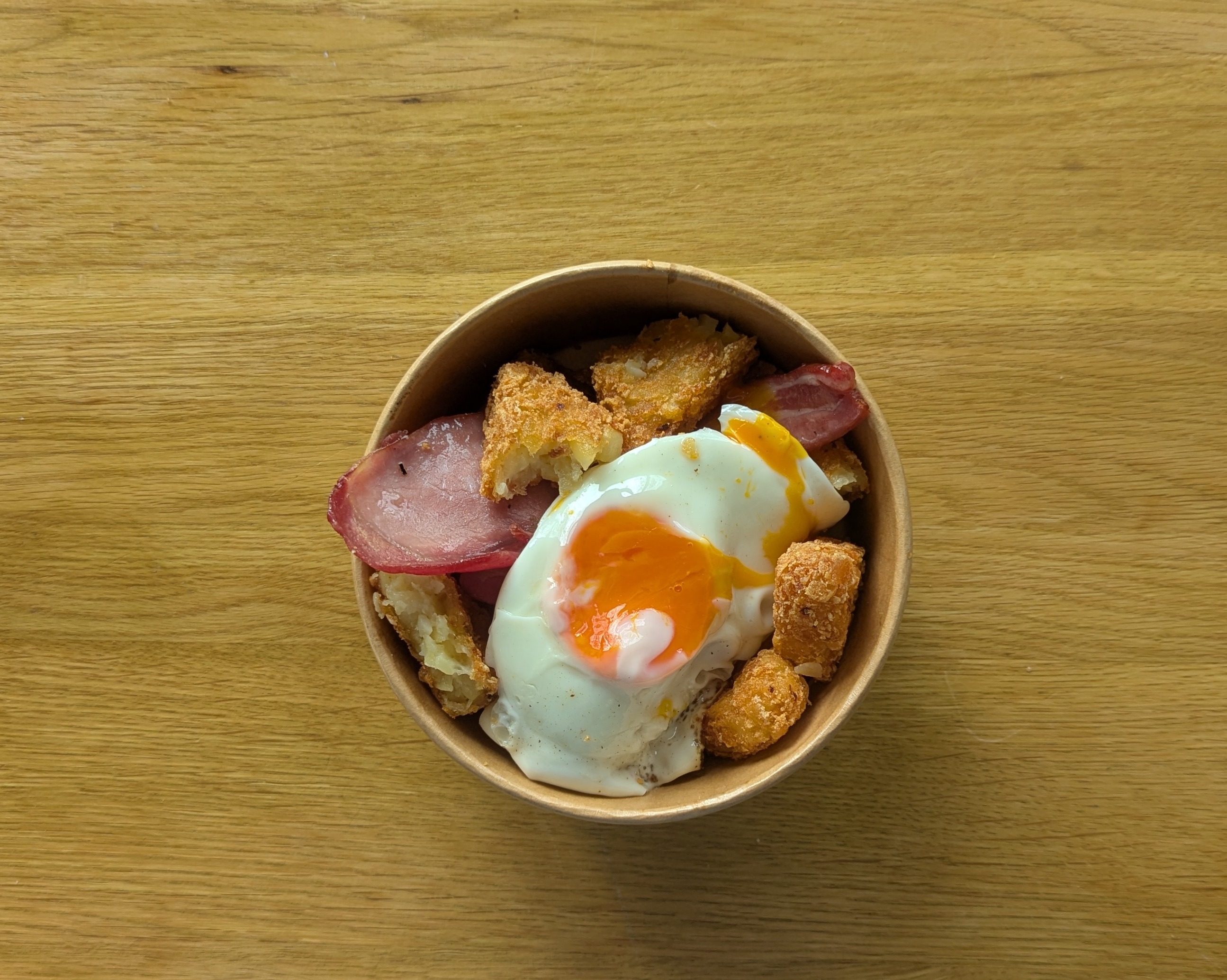 A rustic bowl holds golden tater tots, crispy bacon, and a perfectly poached egg with a vibrant orange yolk, set against wood grain.