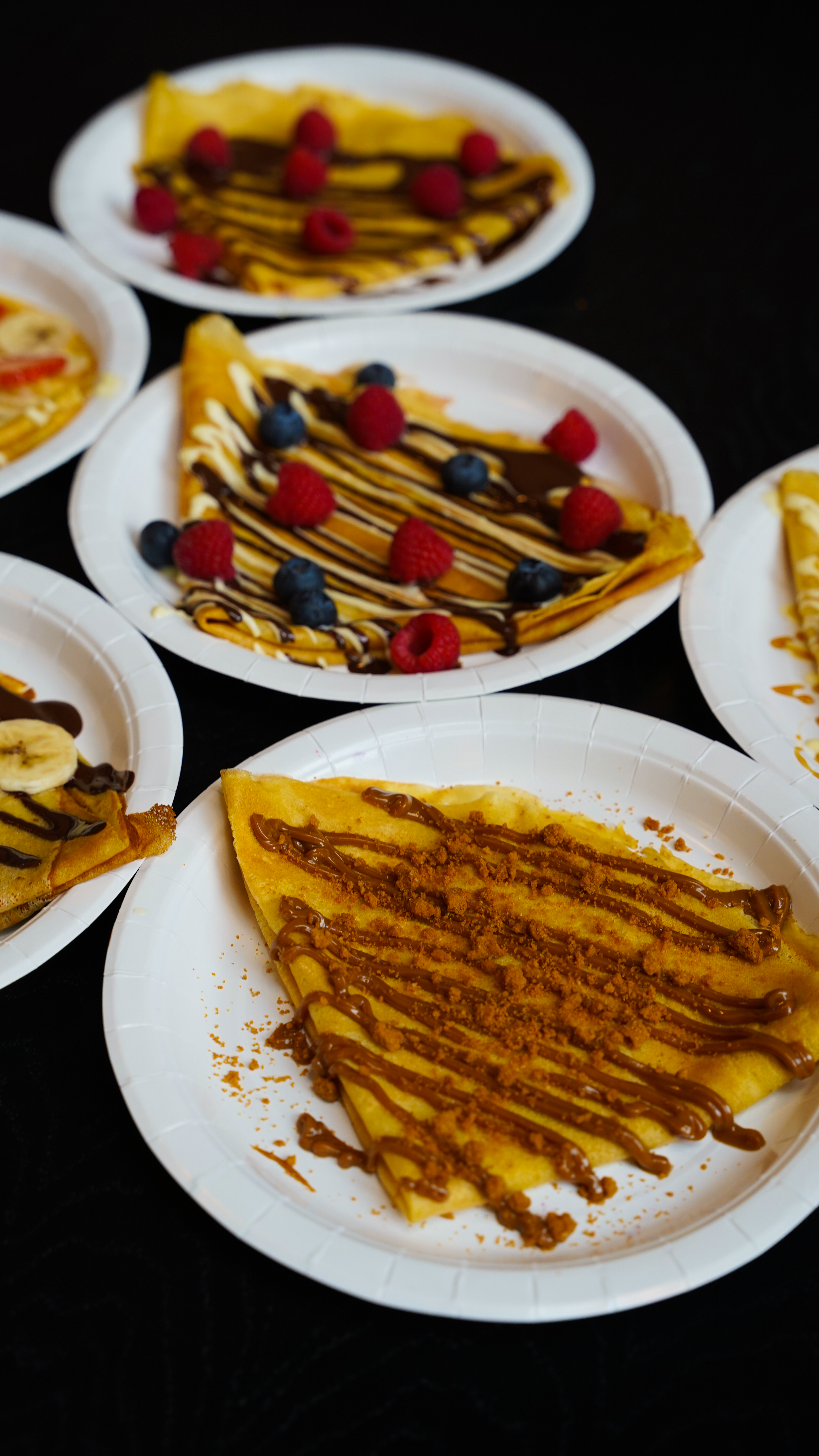 Colorful crepes are artfully arranged on white plates, topped with fresh raspberries, blueberries, and drizzled with caramel sauce.