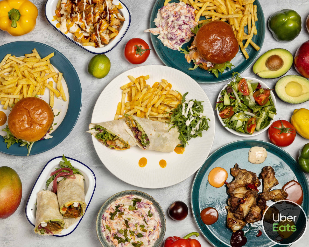 Overhead catering spread of burgers, wraps, fries, salads, avocado, and grilled chicken on teal and white plates, bright studio-lit.