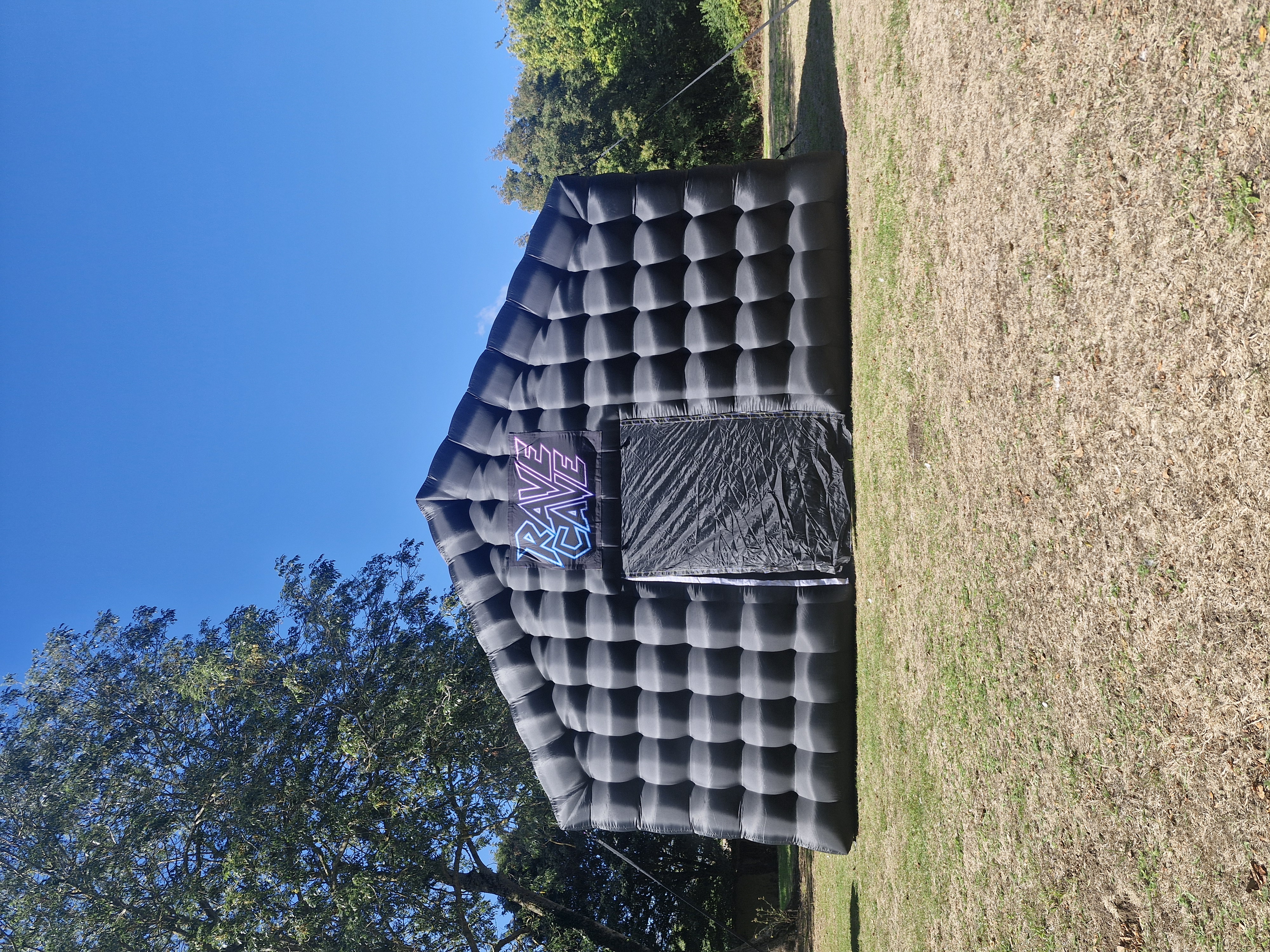 Black inflatable Rave Cave tent on grass, centered under a bright blue sky, with neon sign and crisp event-rental setup.
