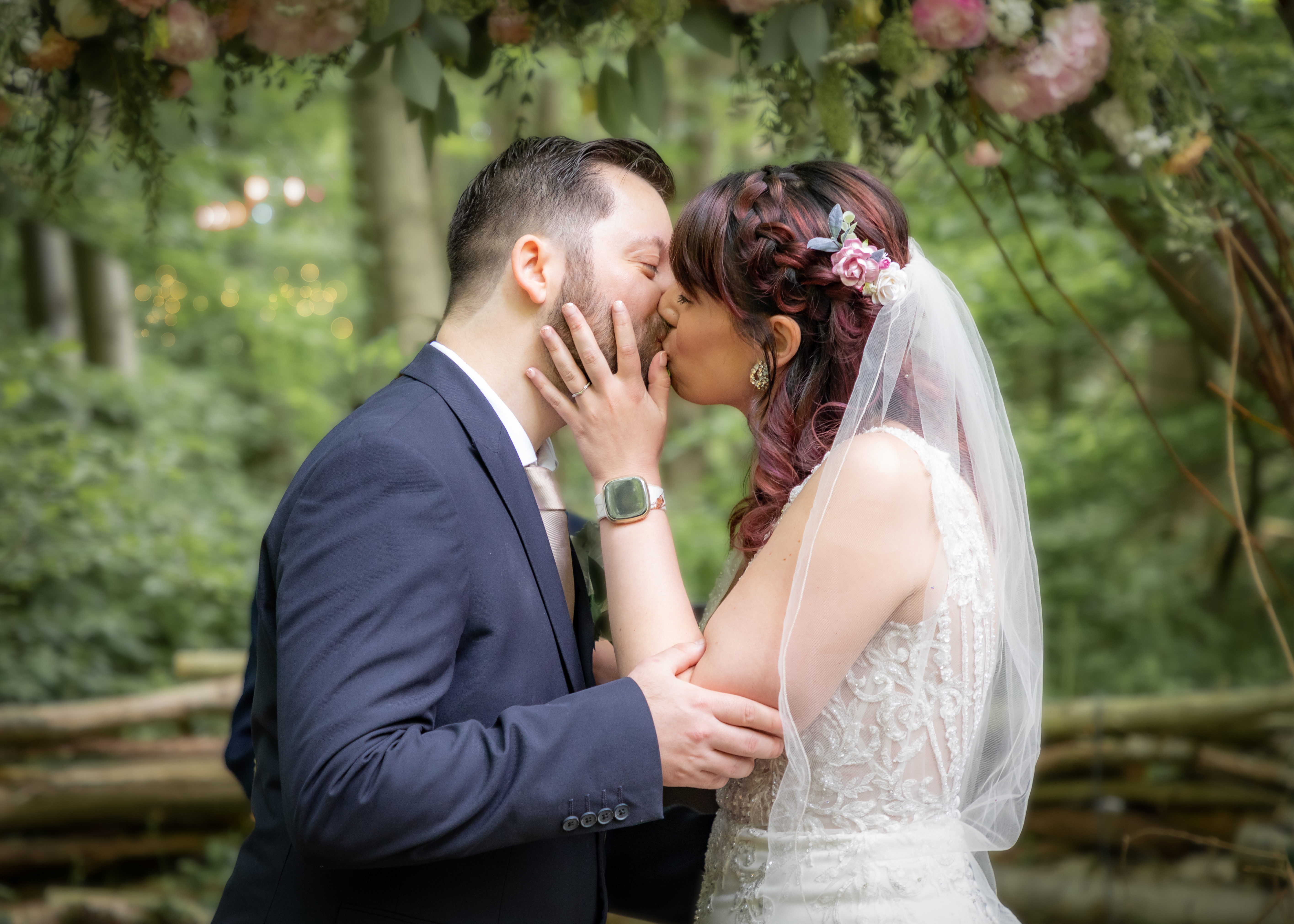 A couple kisses beneath a floral archway in a lush green forest, surrounded by soft, dappled light, creating a romantic atmosphere.