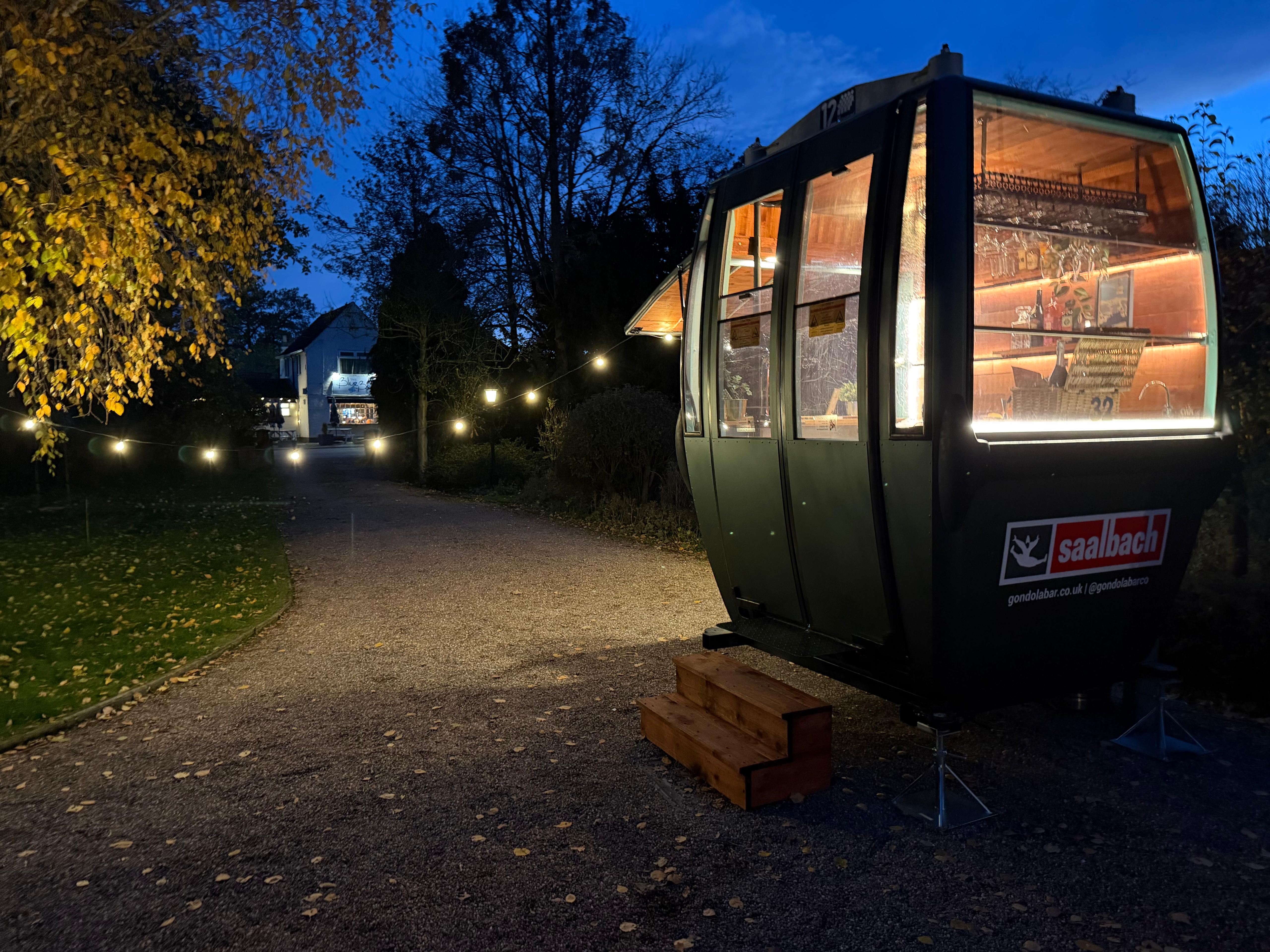 A cozy gondola bar lit by warm, inviting lights showcases glassware and rustic wooden accents, nestled along a gravel path amid autumn leaves.