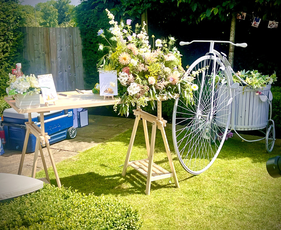 A rustic wooden table displays a vibrant floral arrangement beside a vintage bicycle, with bright sunlight illuminating the inviting garden setup.