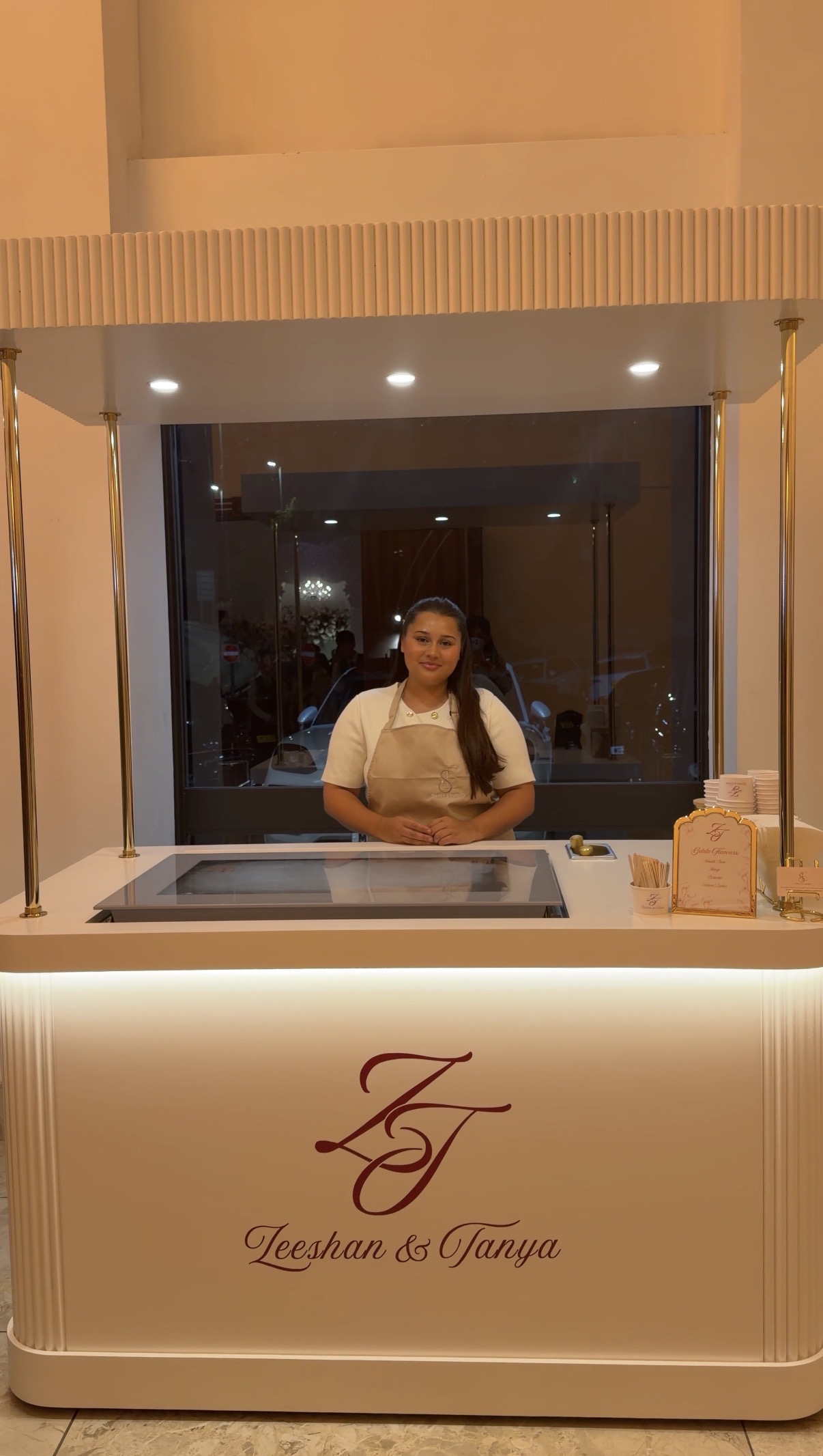 Elegant gelato catering counter with gold posts, white marble finish, branded signage, and warm evening lighting, manned by a smiling attendant.