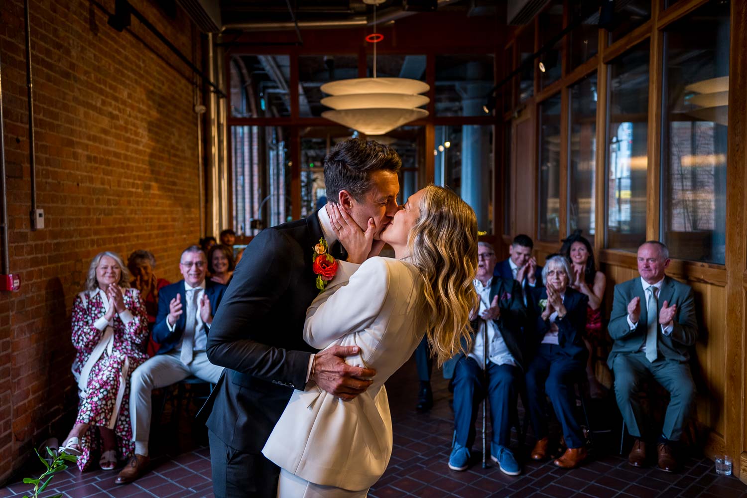 A joyous couple kisses in a cozy venue with exposed brick walls and warm overhead lighting, surrounded by applauding guests in elegant attire.