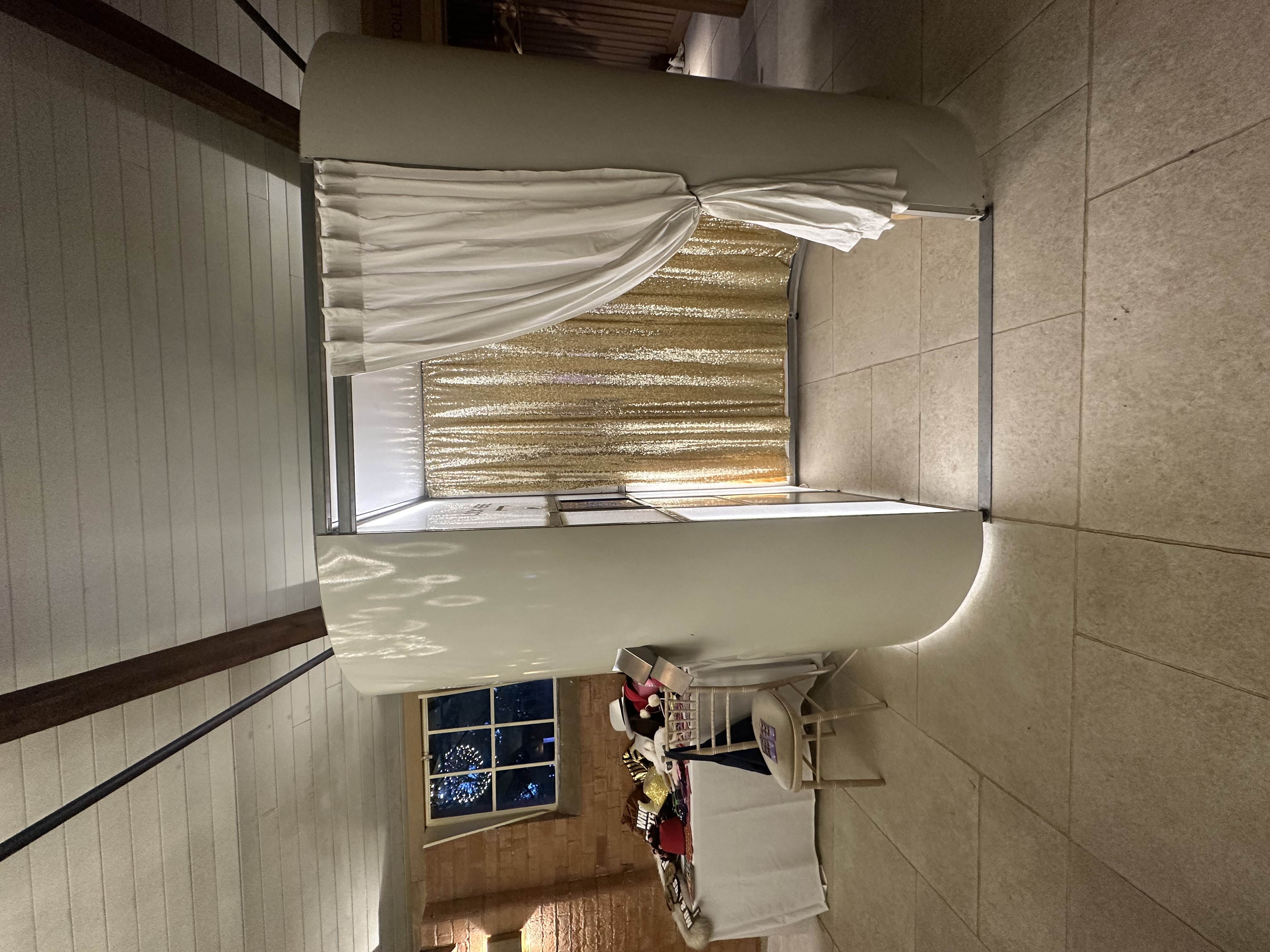 Elegant photo booth with gold sequin backdrop, white drapes, soft LED glow, and a nearby prop table in warm event-catering ambiance.