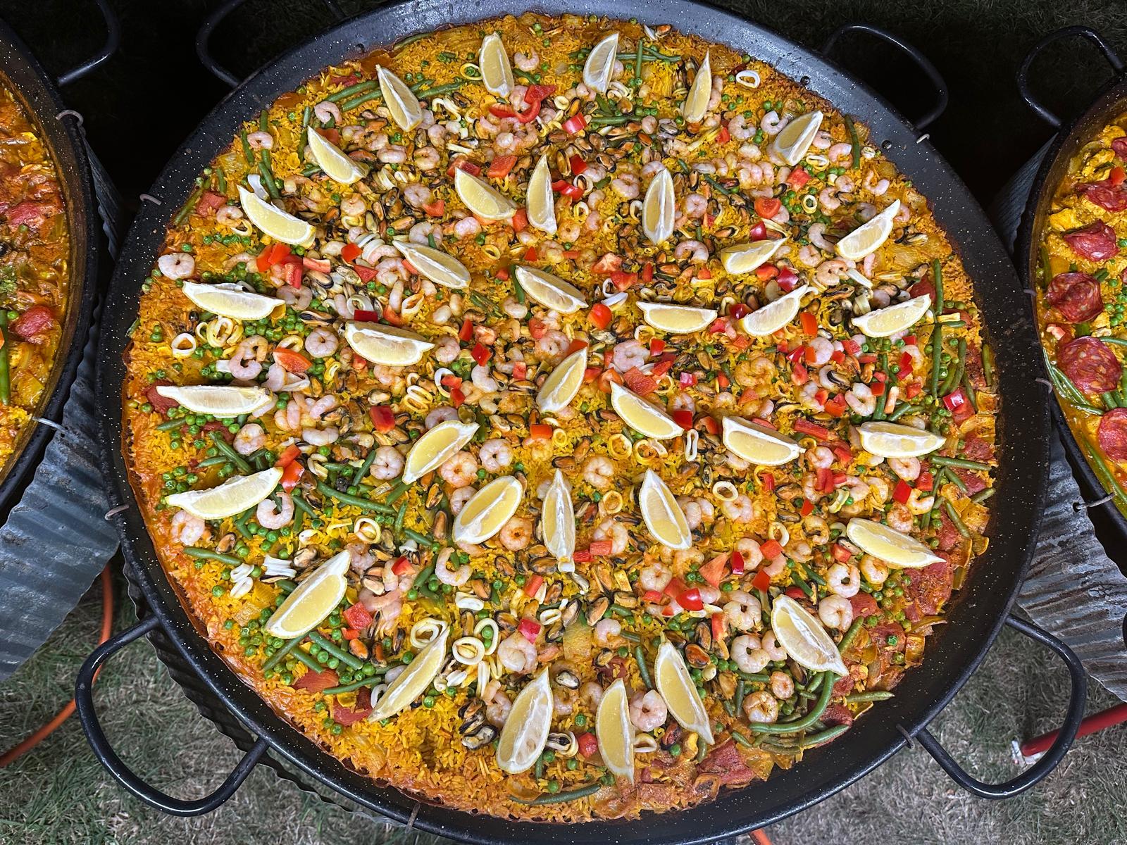 Giant seafood paella in a black catering pan, topped with shrimp, mussels, lemon wedges, peppers, and green beans under bright daylight.