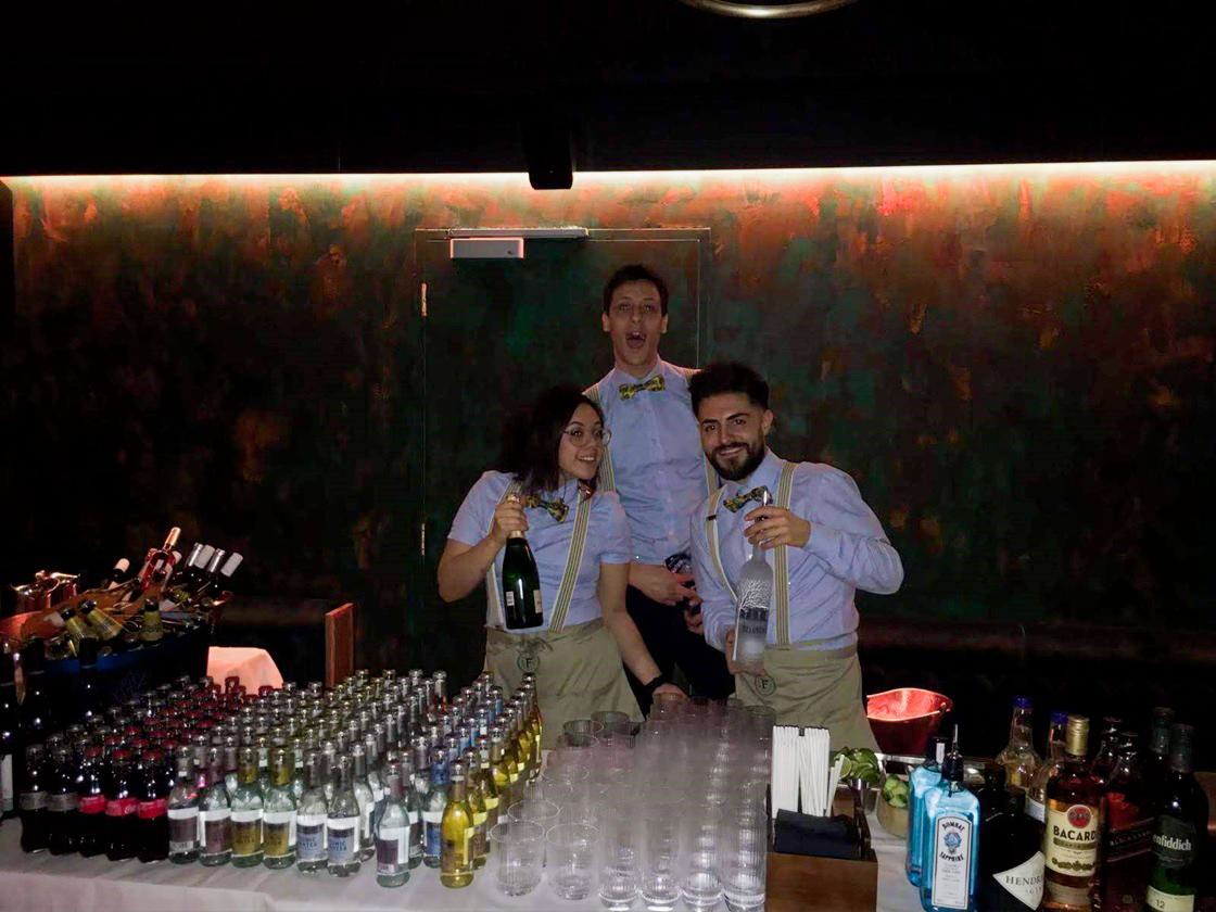 Three cheerful bartenders in light blue shirts and bow ties serve drinks at a well-stocked bar with sparkling glassware, enhancing the festive vibe.