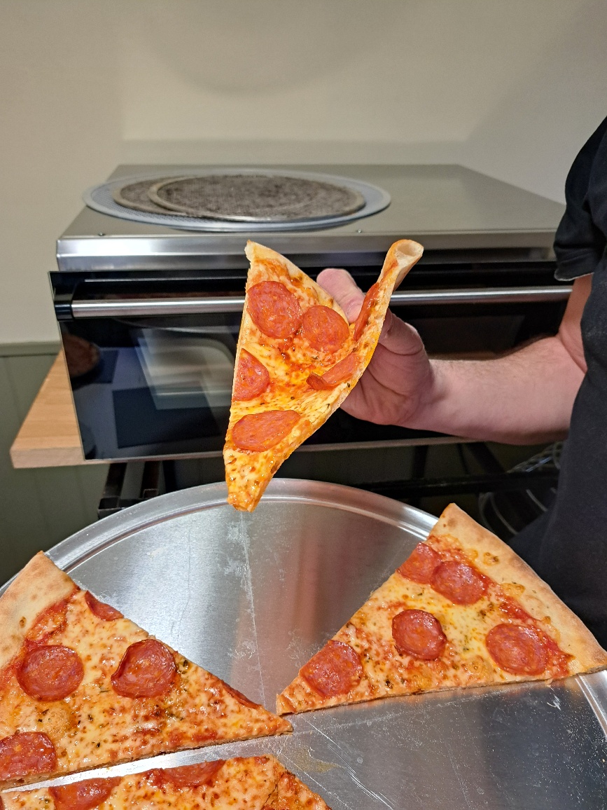 A hand lifts a slice of pepperoni pizza from a silver round tray, with melted cheese glistening under warm kitchen lights, showcasing freshness.