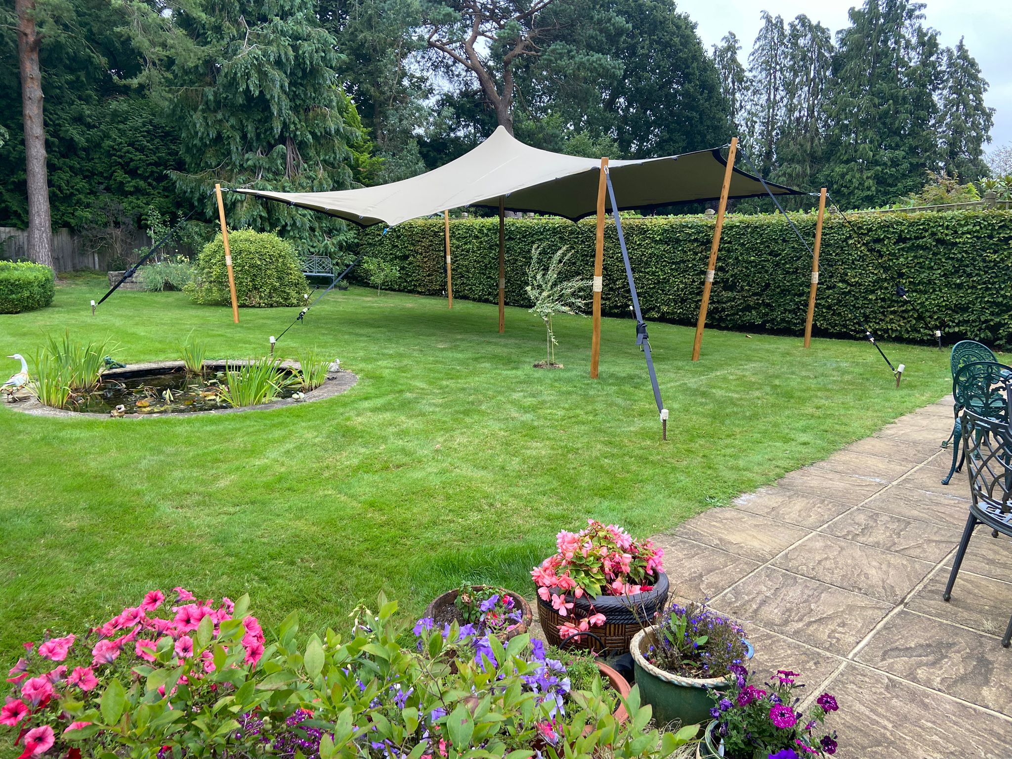 A serene garden features a large beige canopy held by wooden poles, vibrant flower pots, and a tranquil pond, all under soft, natural light.