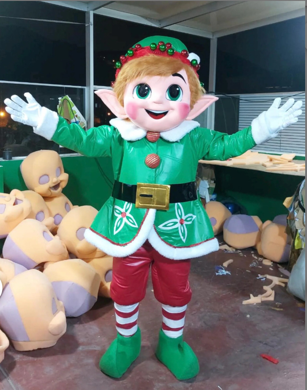 A cheerful elf in a vibrant green outfit with red accents and a big smile stands in a workshop surrounded by unfinished decorative heads, creating a playful and festive atmosphere.