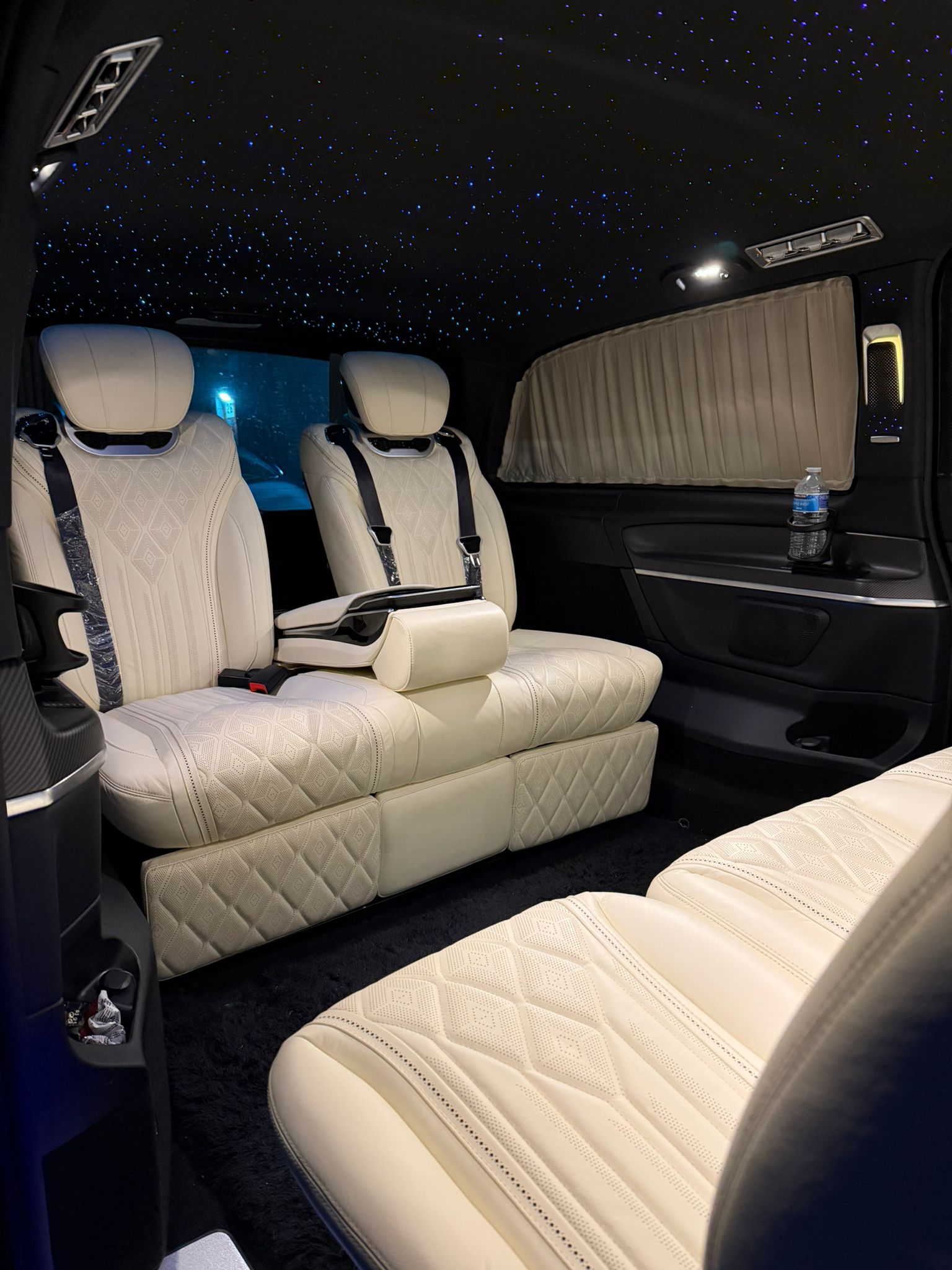 Luxury chauffeur van interior with cream quilted leather captain seats, starry LED ceiling, beige curtains, and polished black trim.