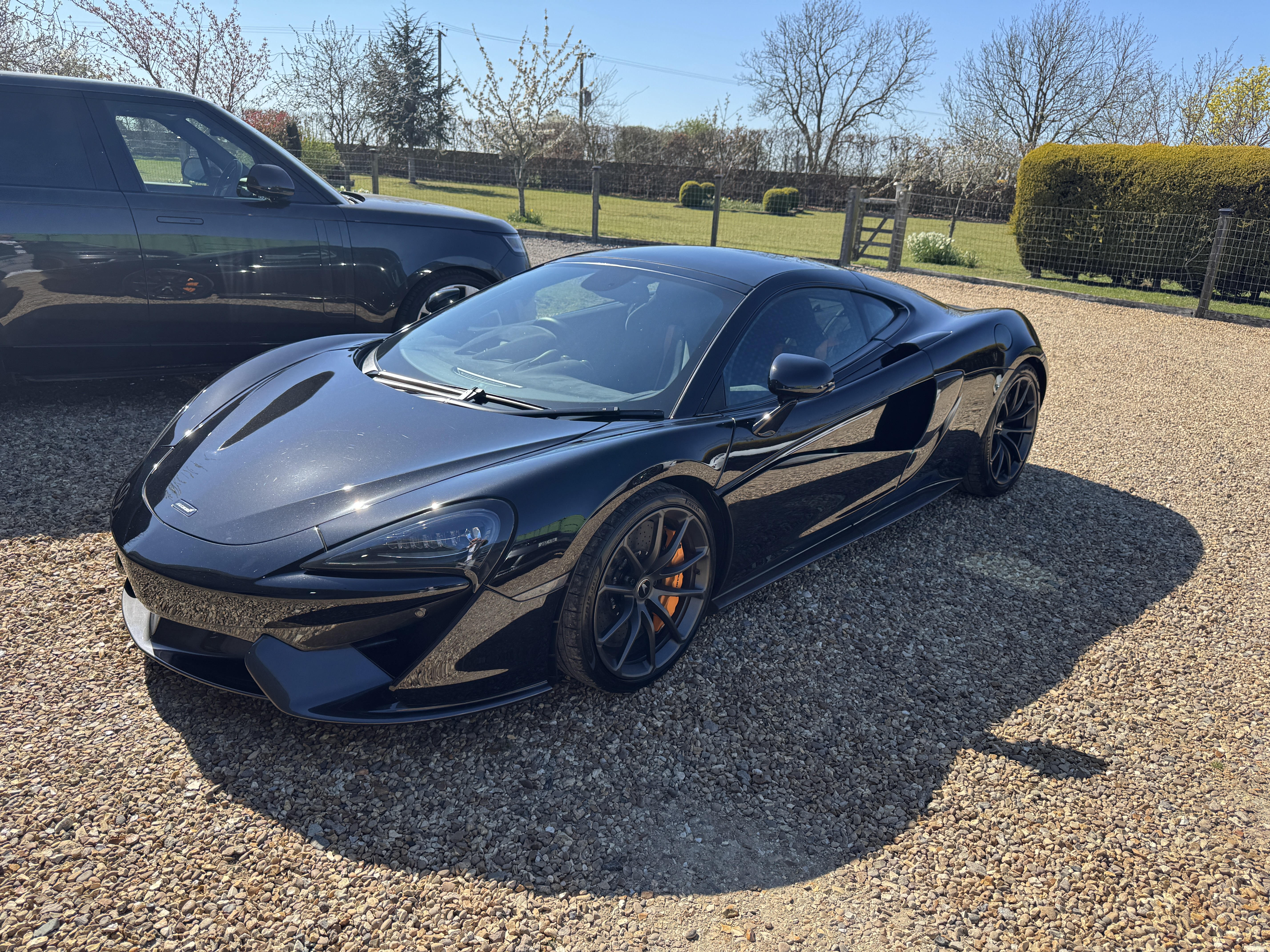 A sleek black McLaren sports car gleams under bright sunlight, parked on textured gravel beside a luxurious SUV, showcasing elegance.