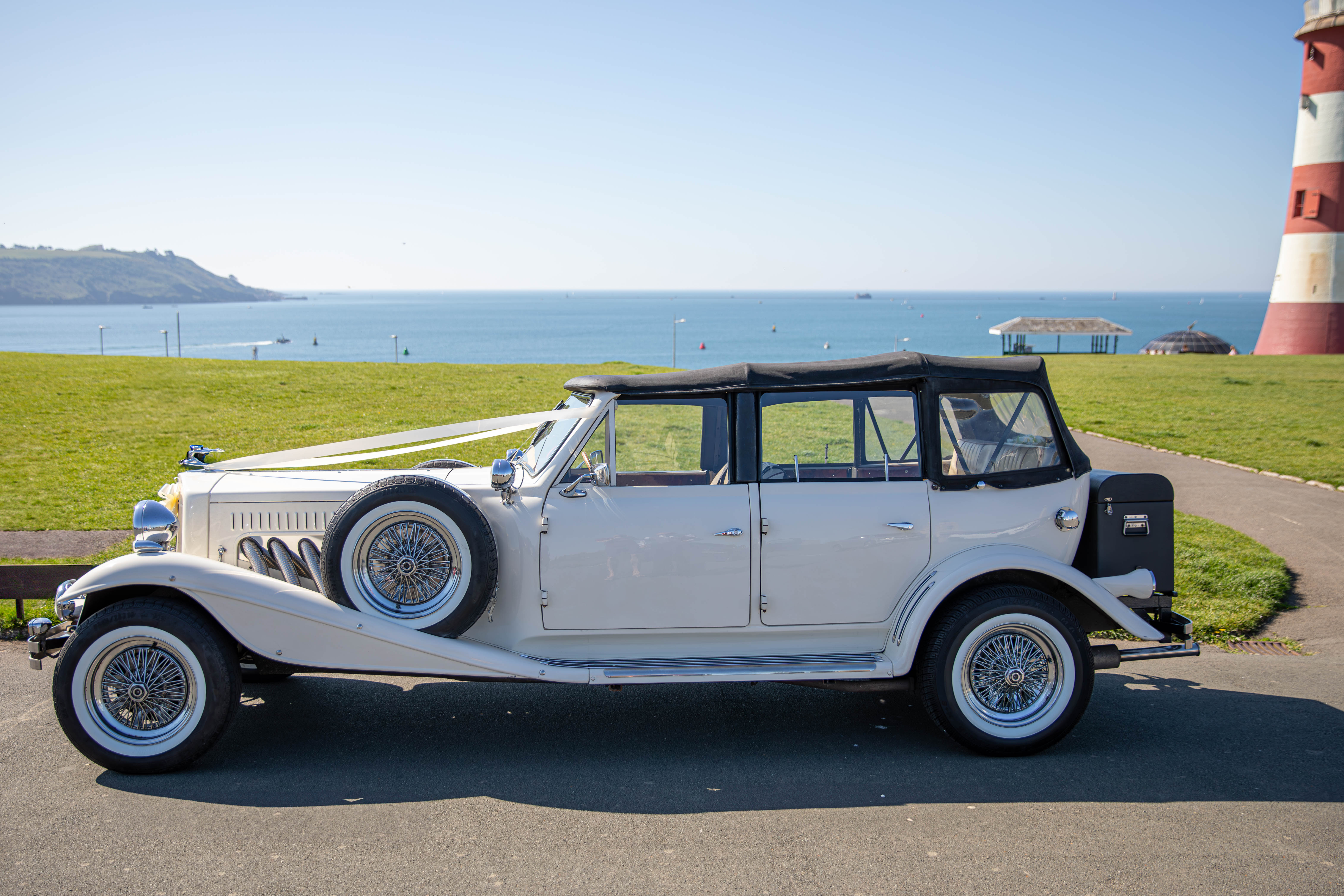 A classic white vintage car, adorned with a satin ribbon, is parked against a serene seaside backdrop, showcasing polished chrome accents.