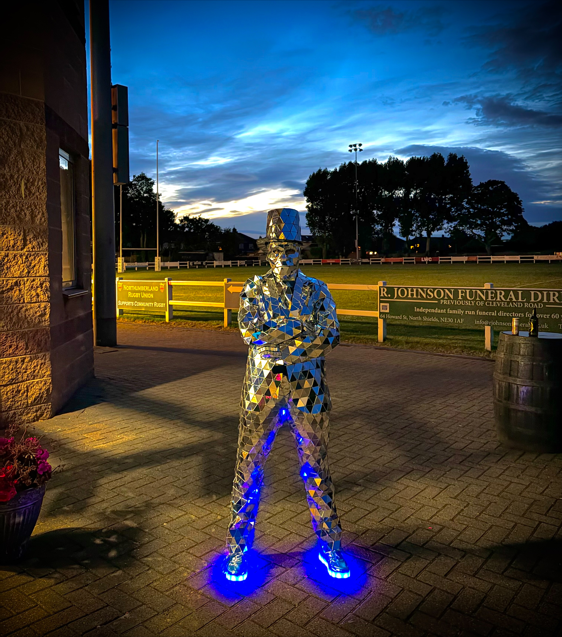 Mirrored silver human sculpture with blue-lit shoes stands on brick paving beside rugby-field signage at dusk, creating a striking event-installation mood.