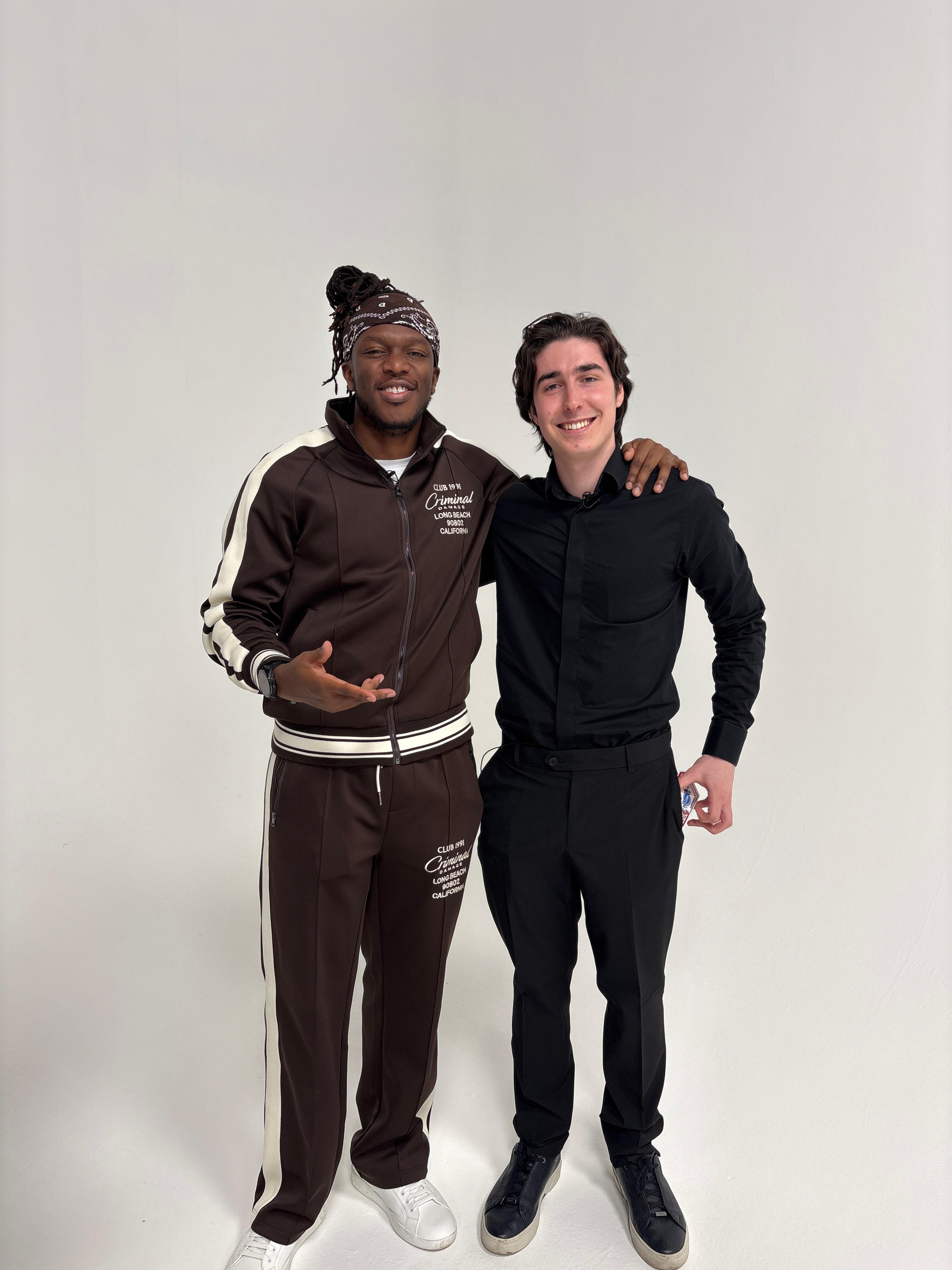 Two smiling men pose arm-in-arm against a clean white studio backdrop, wearing black and brown casual outfits under bright, even lighting.