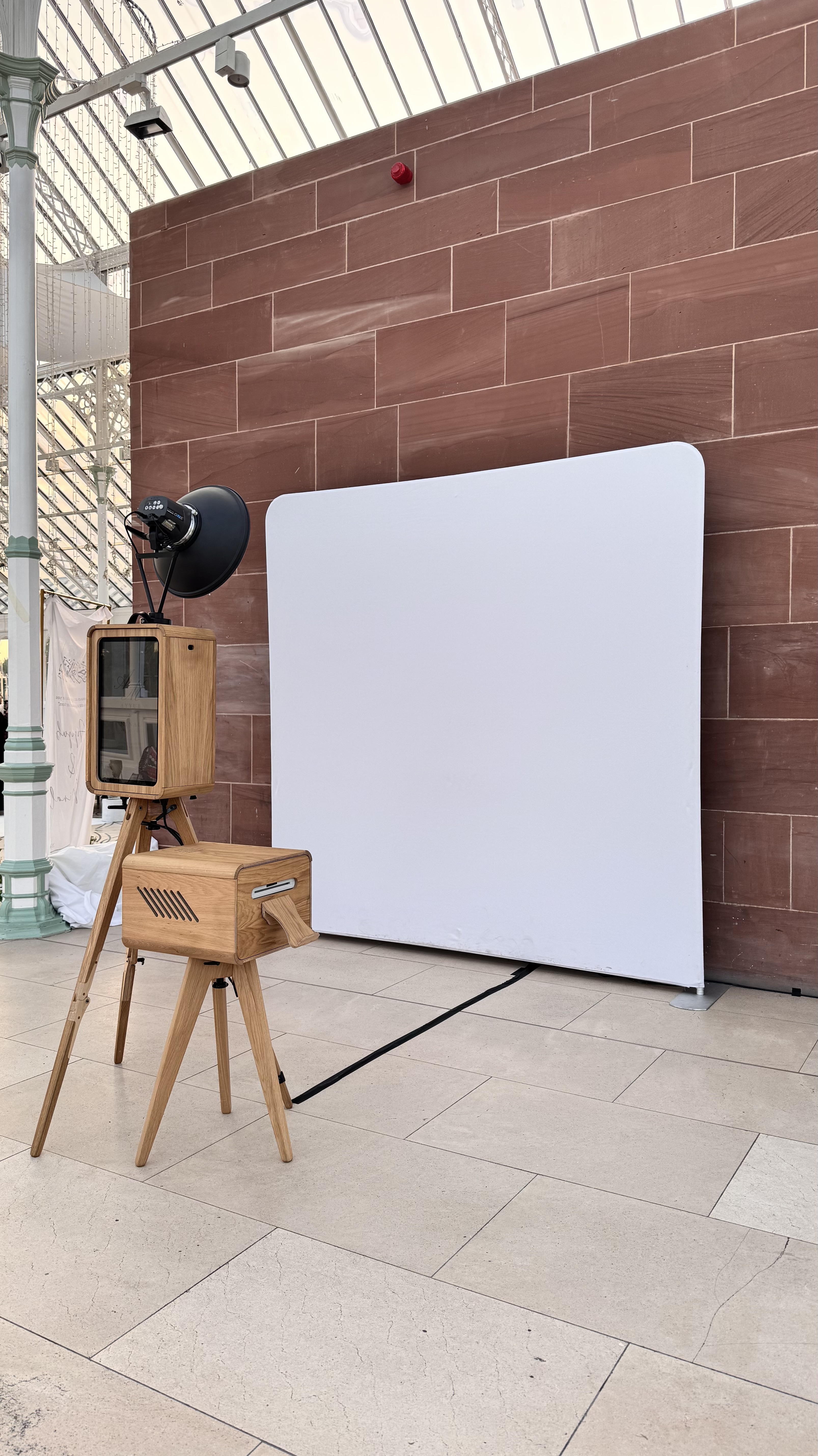 A stylish wooden vintage photo booth with an antique projector and a sleek white backdrop is elegantly arranged on polished stone tiles, illuminated by natural light.