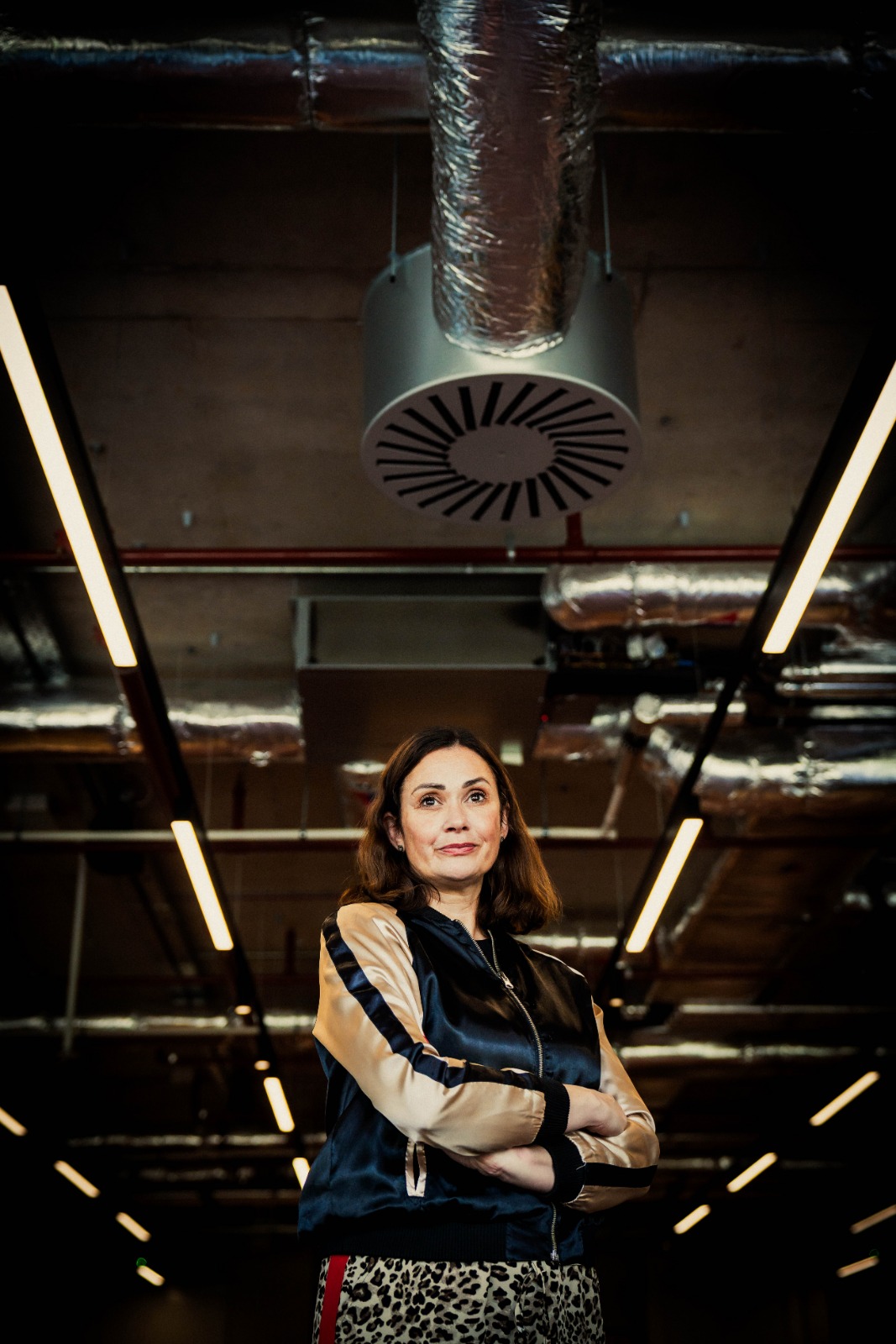Confident woman in satin bomber jacket stands under industrial ductwork and linear lights, creating a sleek, modern catering venue vibe.