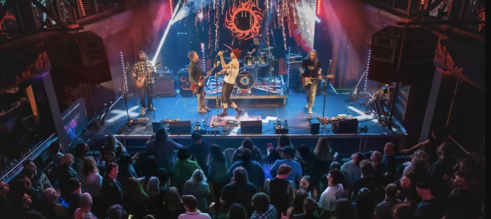 A lively band performs on stage under vibrant lighting, with a crowd swaying and cheering, creating an energetic, spirited atmosphere.