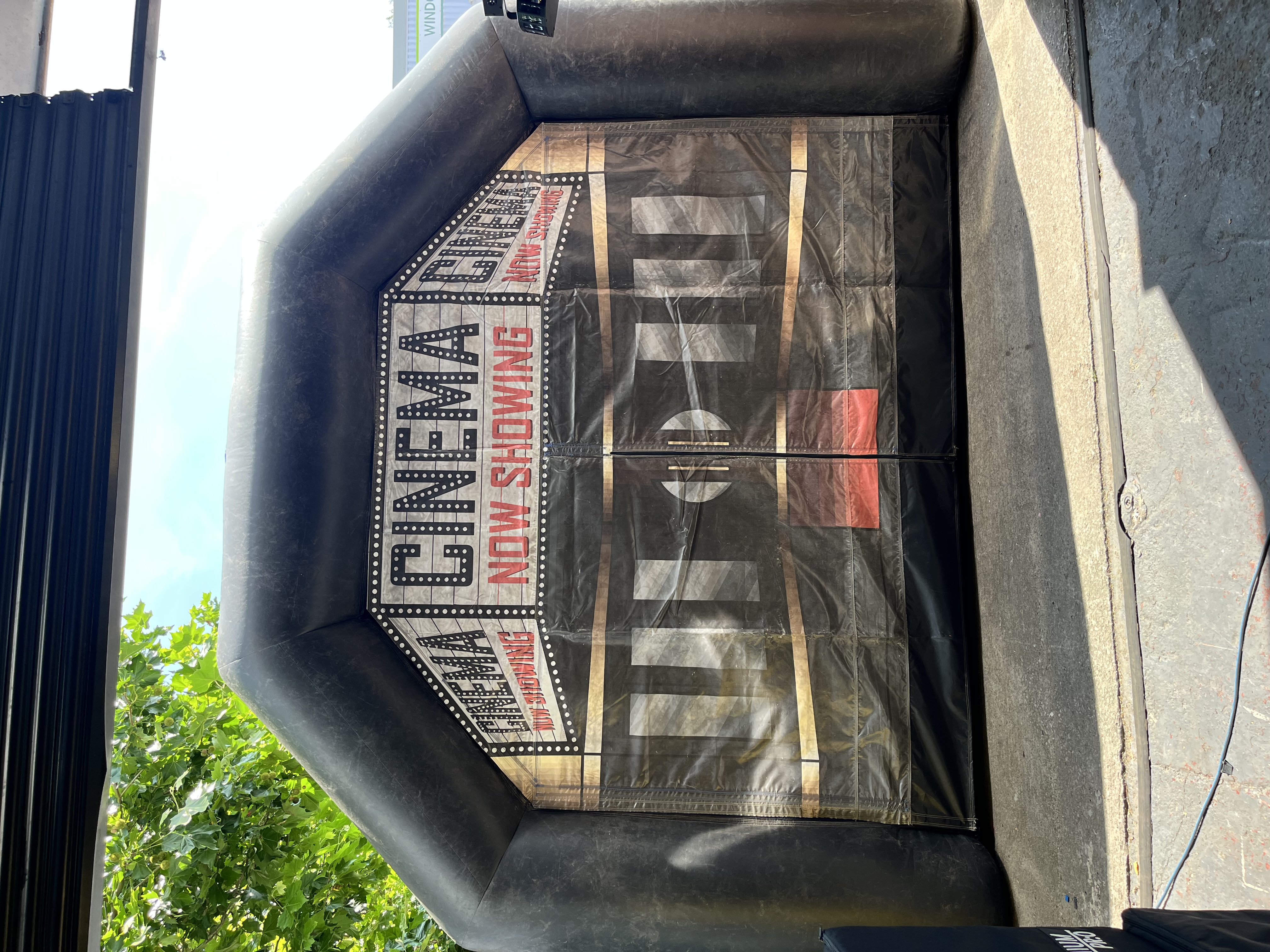 Black inflatable cinema entrance with marquee-style “NOW SHOWING” signage, set outdoors in bright daylight for event catering or movie nights.