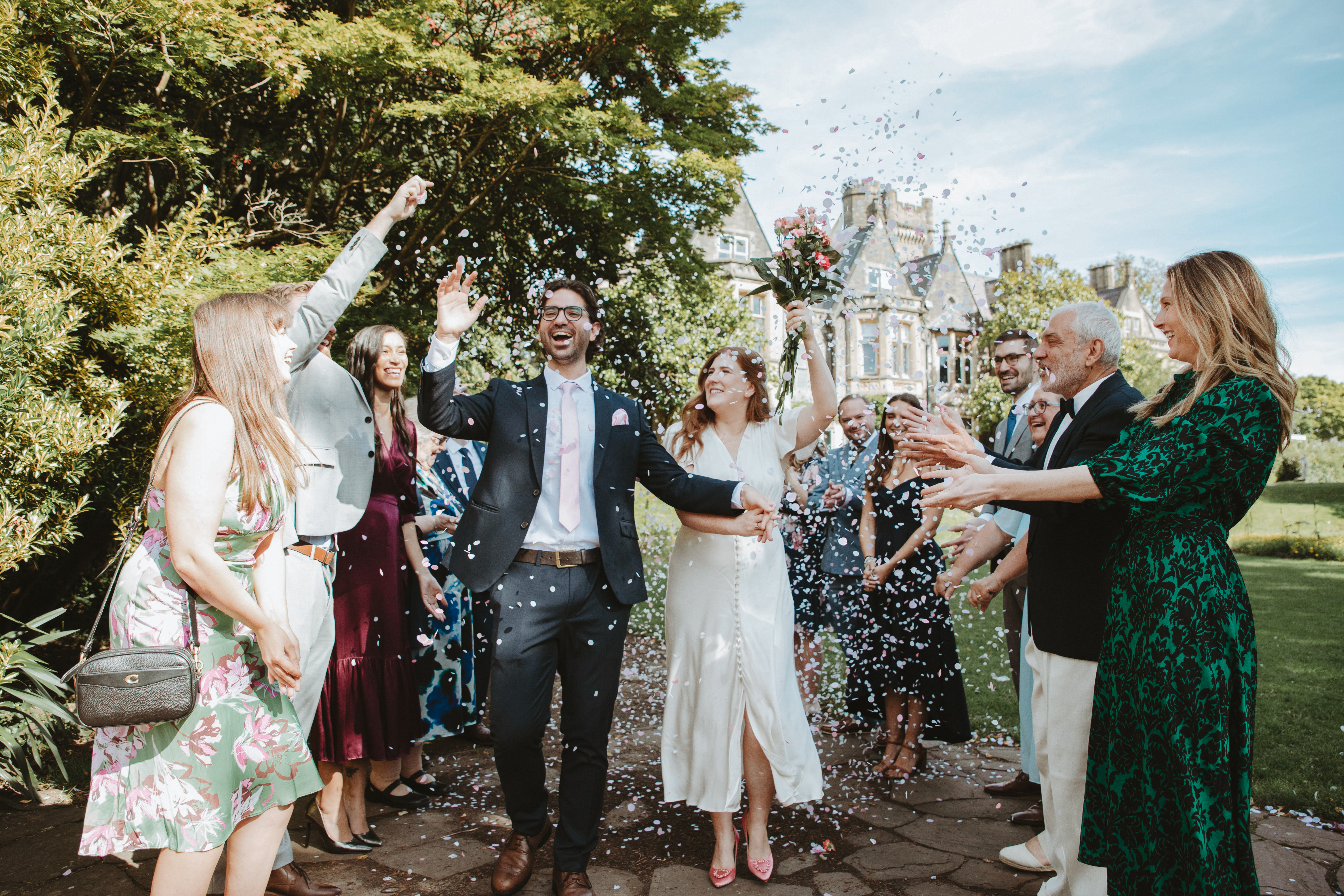 A joyful wedding celebration outdoors, with guests in stylish attire throwing colorful confetti around a couple wearing elegant outfits.