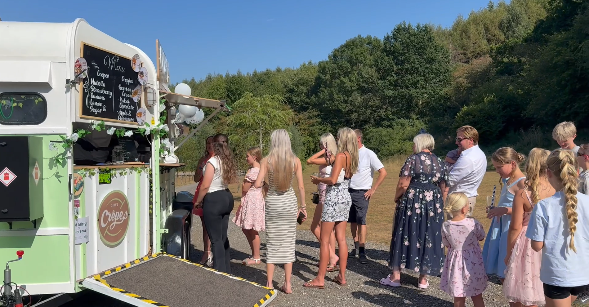 Guests queue at a mint-green vintage crepe van, with chalkboard menu and bright balloon accents, in sunny outdoor catering.