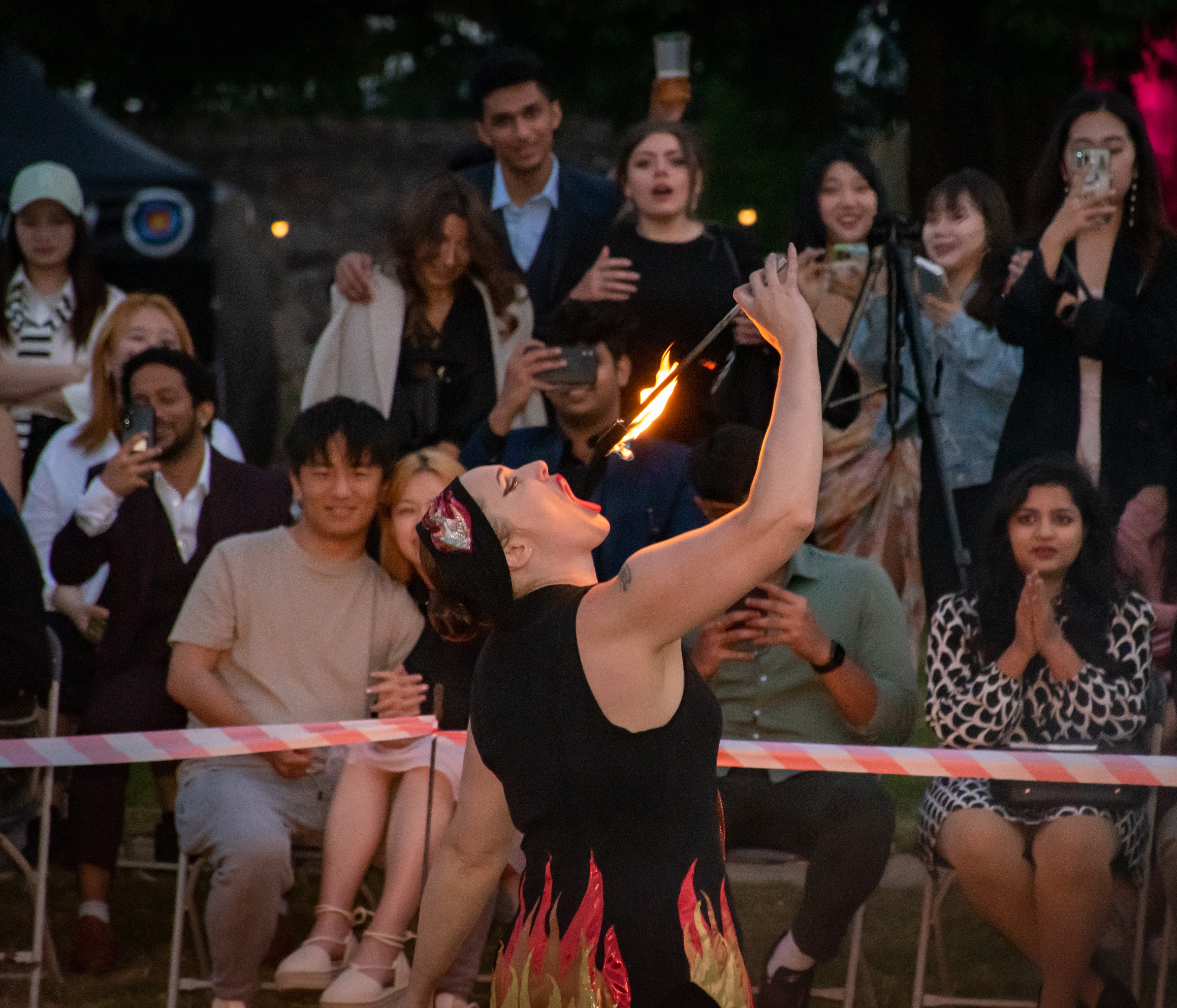 A fire performer captivates a crowd under soft evening lights, creating an electric atmosphere with vibrant flames against attentive faces.