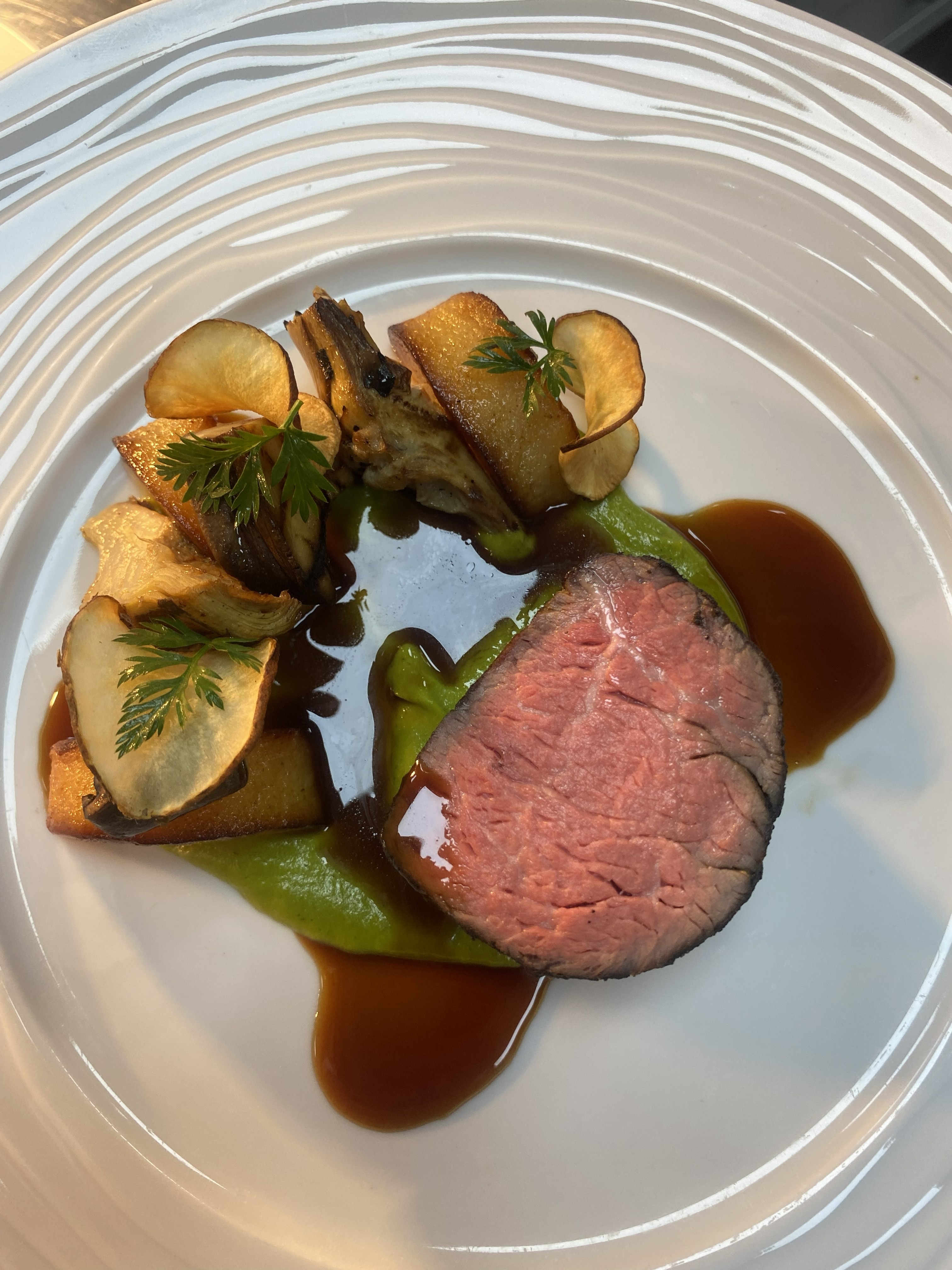 A beautifully plated dish features tender pink beef surrounded by vibrant green puree, roasted potatoes, and delicate garnishes under soft, ambient lighting.
