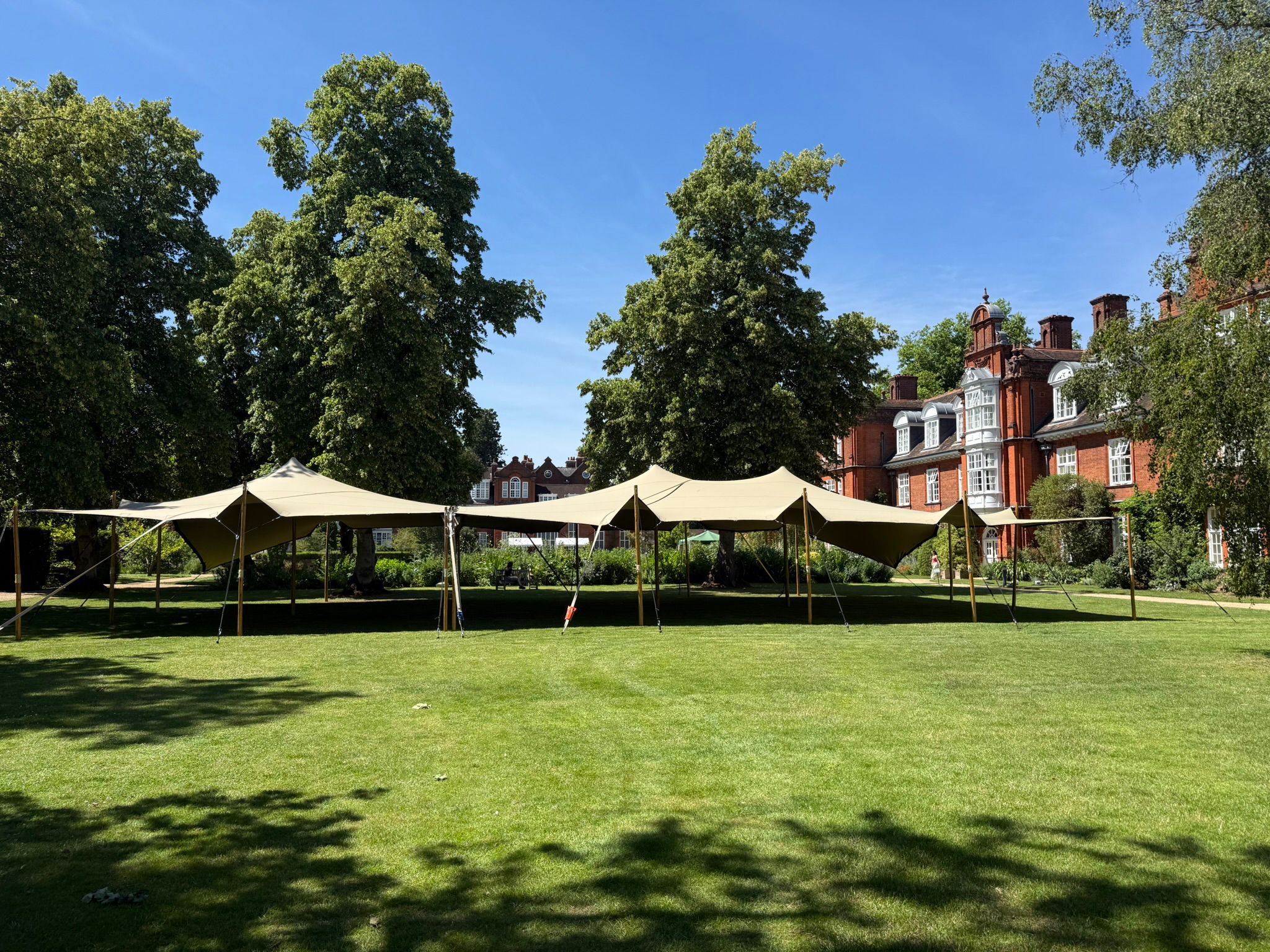 Beige event canopies stretch across a manicured lawn beside a red-brick manor, under bright daylight with clean catering setup vibes.
