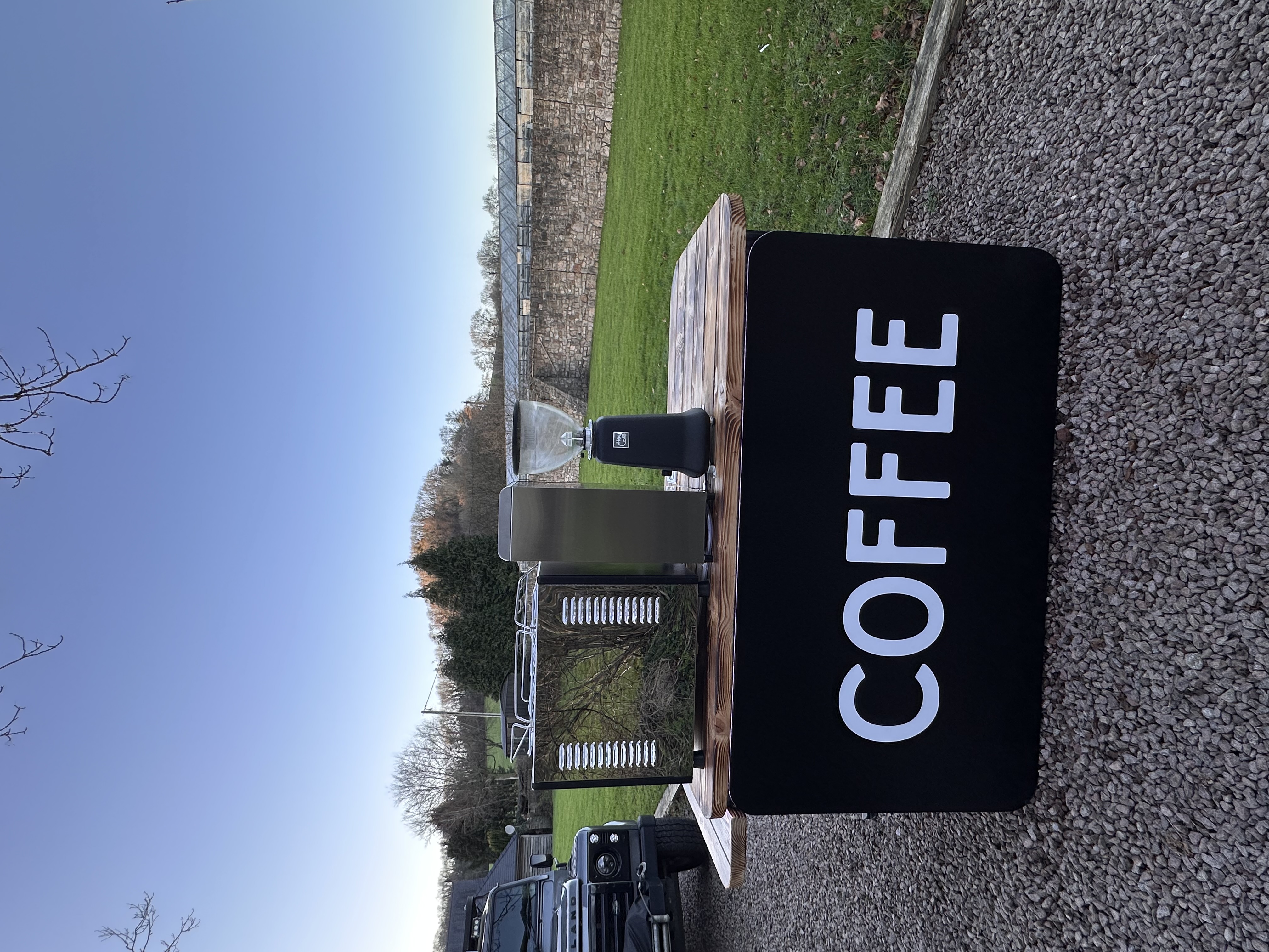 A rustic wooden coffee stand displays sleek equipment, including a grinder and brewer, with a sunny green lawn backdrop, hinting at quality catering.